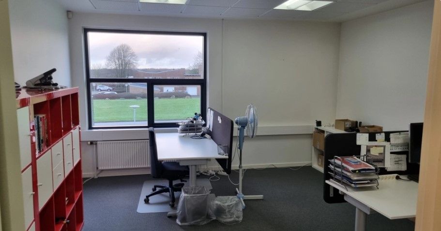 Kontor med bord, stol, ventil, fan og bibliotek med bøger samt udsigt til græsplæne og træer