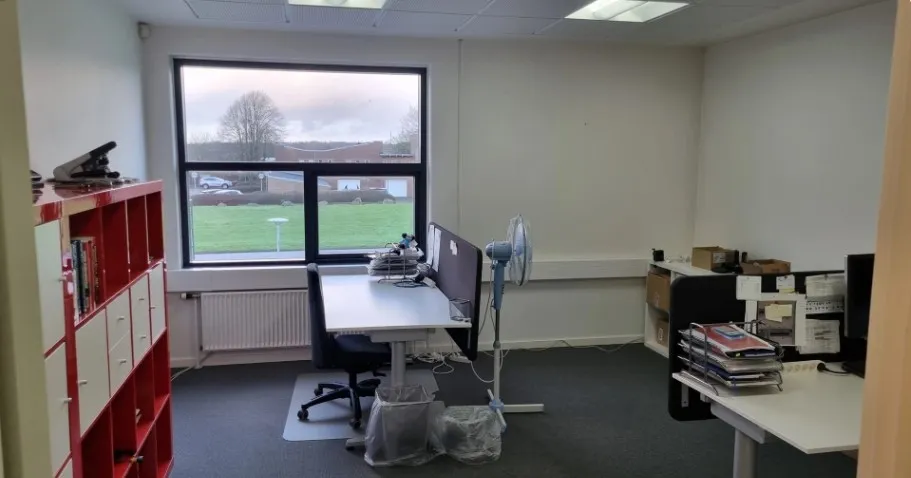 Kontor med bord, stol, ventil, fan og bibliotek med bøger samt udsigt til græsplæne og træer