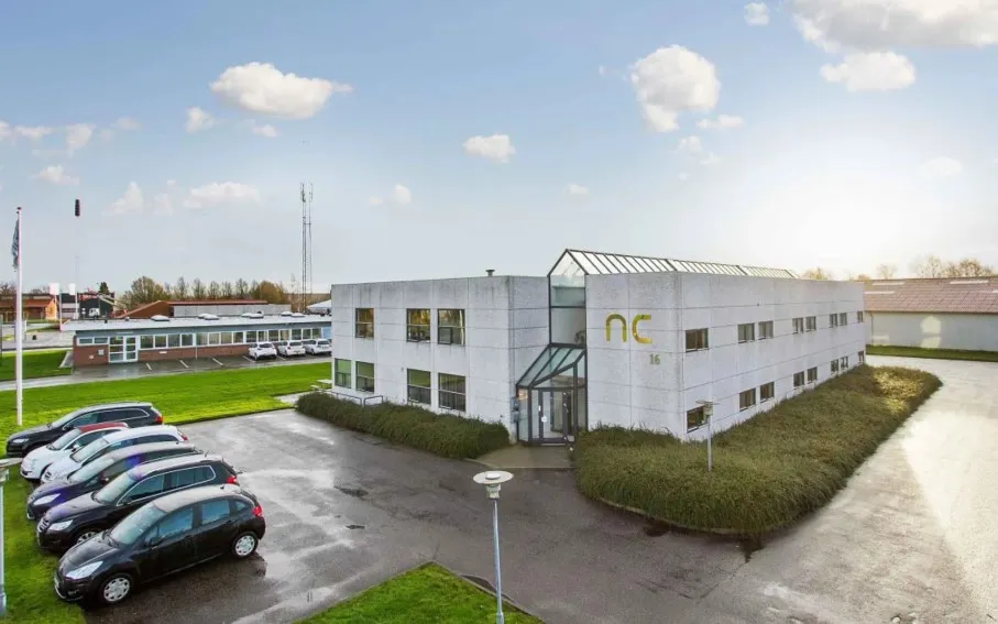 Kontorbygning med NC 16 på facaden og flere parkerede biler foran og bagved bygningen