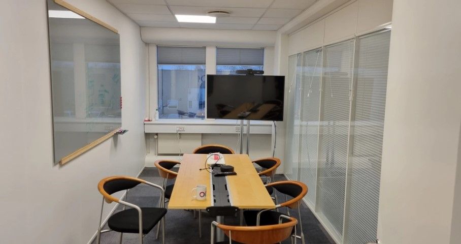 Foto af et kontor med lyse vægge, et gult bord, en tv-skærm og seks stolpestole. Et skrivebord med skrivemaskine og kop står