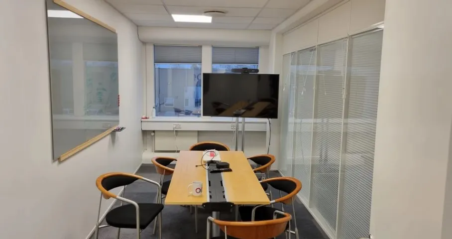 Foto af et kontor med lyse vægge, et gult bord, en tv-skærm og seks stolpestole. Et skrivebord med skrivemaskine og kop står