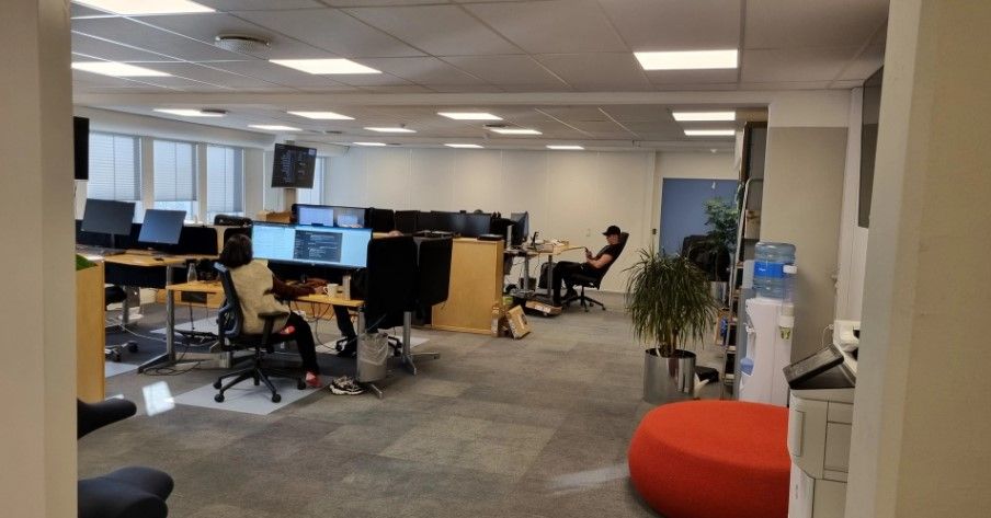 Kontor med flere skriveborde og medarbejdere i en åben stue, med skærme og en gulv med lysegule kvadrater