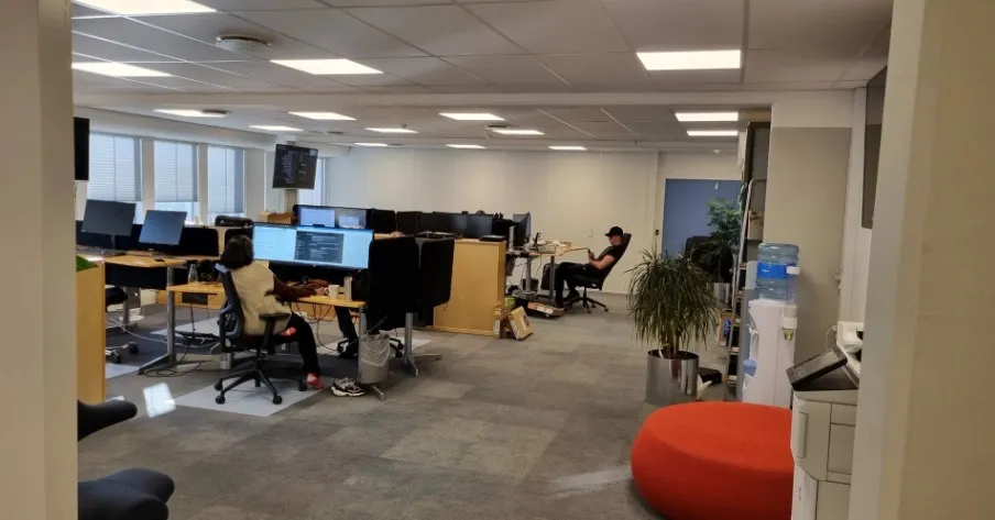 Kontor med flere skriveborde og medarbejdere i en åben stue, med skærme og en gulv med lysegule kvadrater