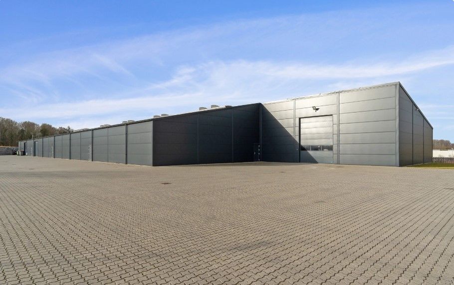 En stor grå industribyggning med brede garagedøre og parkeringsplads. Skyerne i baggrunden er hvide og lyse.