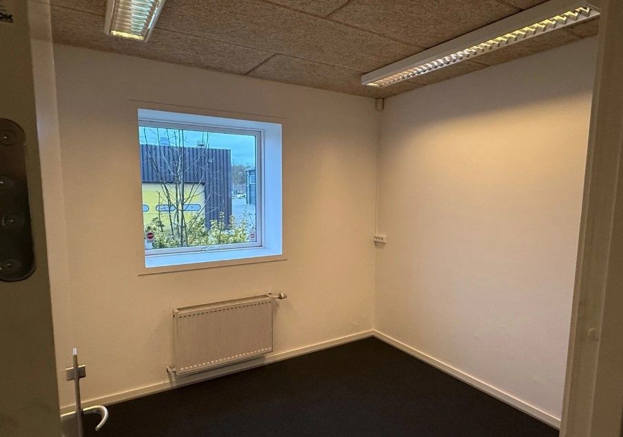 Et tomt kontor med hvide vægge og sort gulv. Vinduet er åbent og der er et radiator under det. Der er to lysekroner på