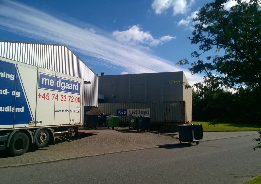 Meldgaard er parkeret foran en industribygning på Kinch Alle 1 i Løsning. Der er mange containere og skraldespande parkeret