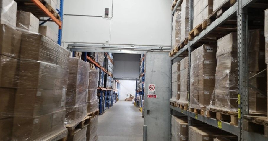 Lager med ekspeditionszone. Rækker med emballage og bokse på lagerhylder. Adgangsdør i bunden og skilt med forbud mod