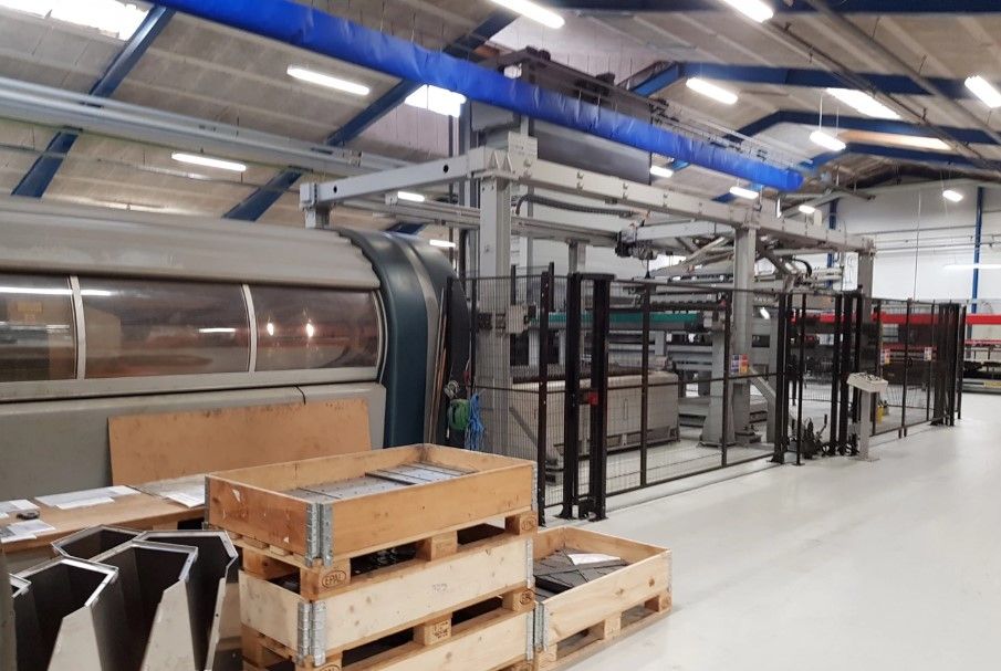 En stor industrihal med flere maskiner, paller, kasser og en stor vægmaske. Et stort stykke pap er pakket på en pallestok.