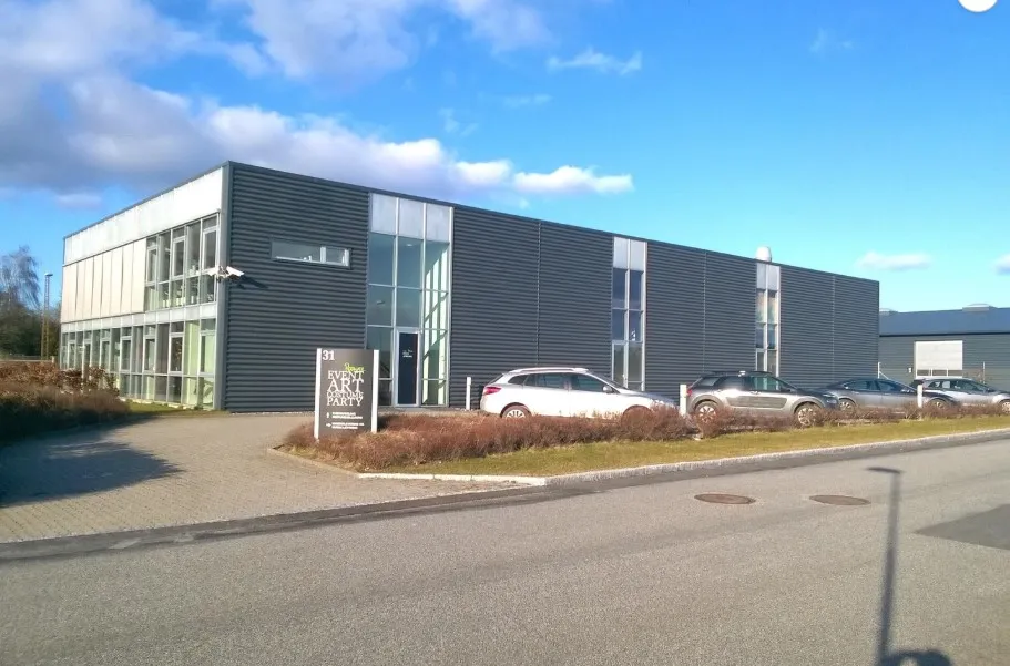 Kontorbyggeri med moderne design og glasvinduer, hvor biler er parkeret foran og skrånende græsplæne med en skilt