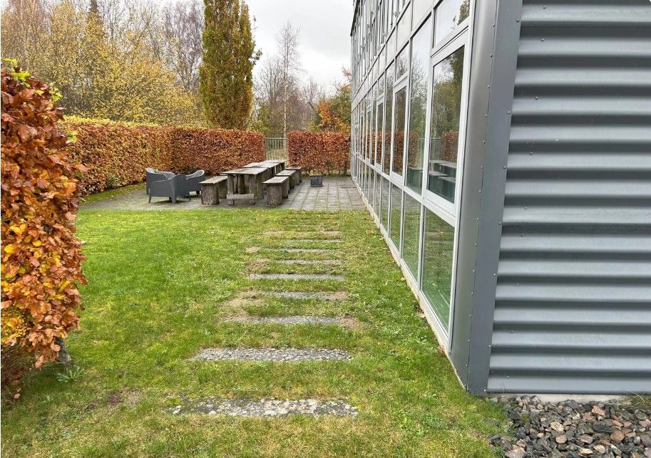 Kontorlokale med en terrasse og stolpeborde og stole. Terrassen er omgivet af grønne hække og græs.