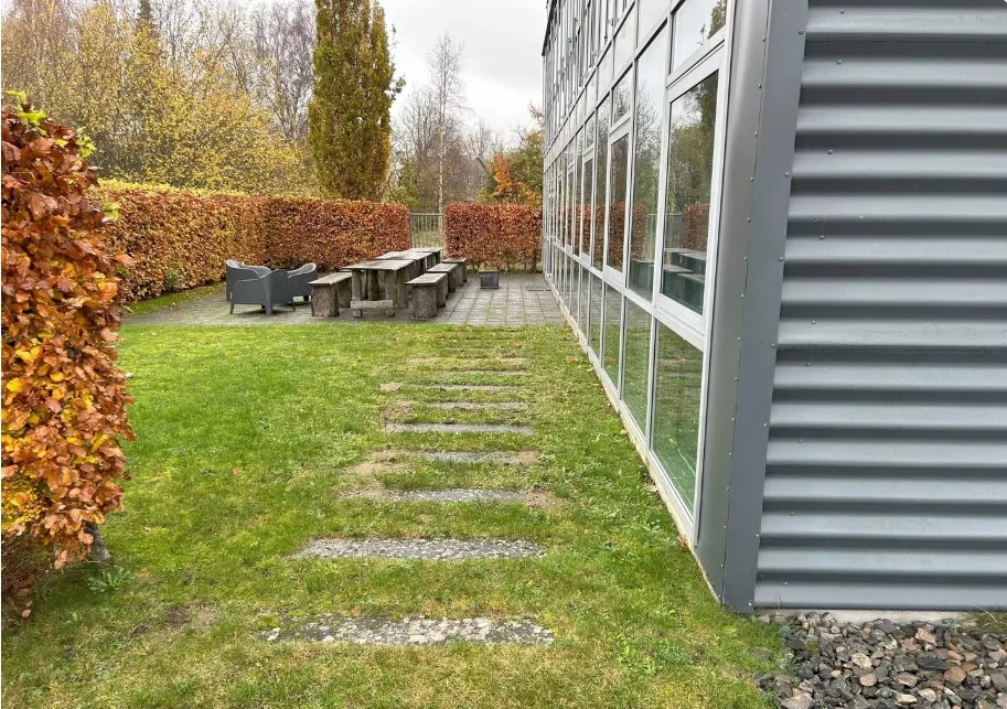 Kontorlokale med en terrasse og stolpeborde og stole. Terrassen er omgivet af grønne hække og græs.