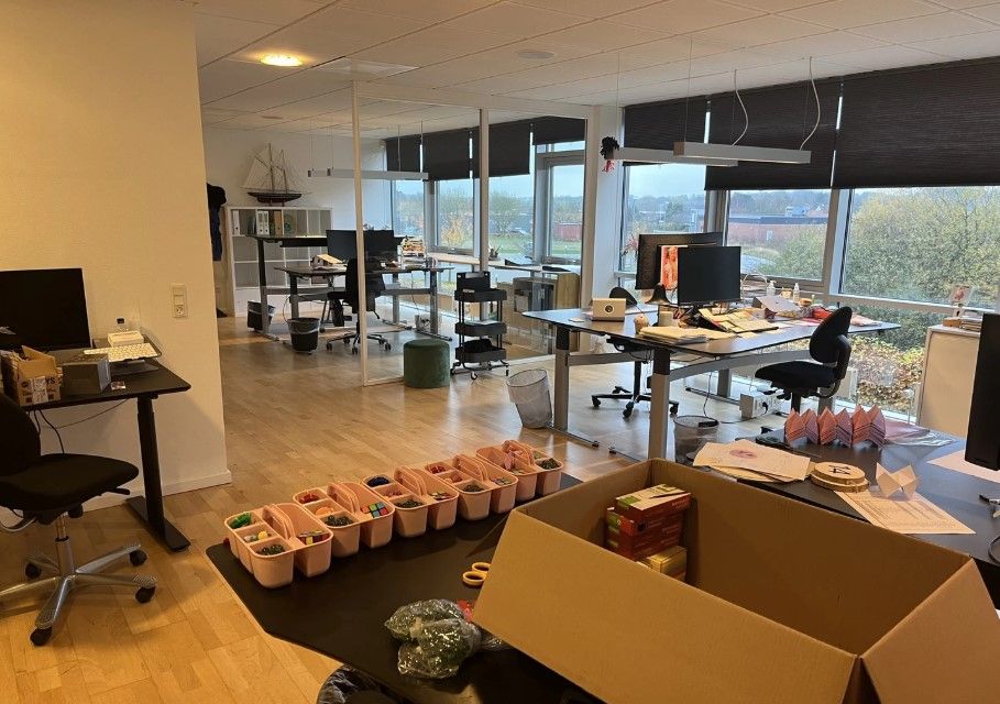 Kontorlokale med skriveborde og stole, papkasser og opbevaringsbakker samt vinduer med udsigt til byen
