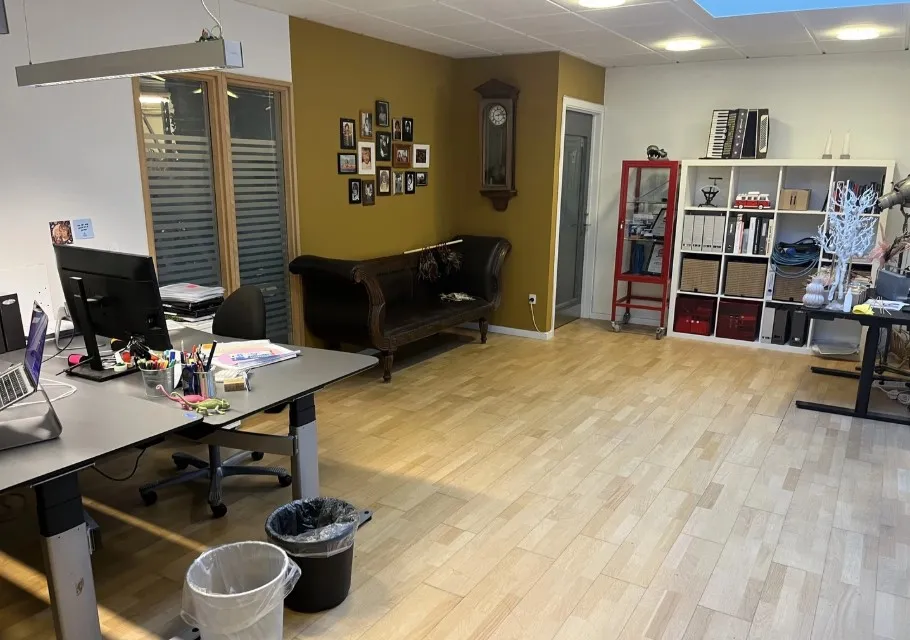 Kontorlokale med skrivebord, computer, skraldespand og lædersofa. Gulv og vægge er lyse og flotte.