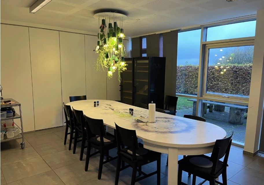 Kontorlokale med en stor rund hvid bordplade, lysekronen med plantegrønt og lamper, træbænke og hegn udenfor vinduet