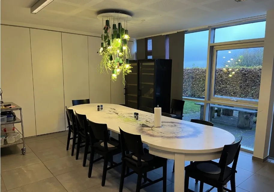 Kontorlokale med en stor rund hvid bordplade, lysekronen med plantegrønt og lamper, træbænke og hegn udenfor vinduet