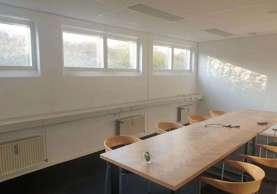 En stor kontorbord, forsynet med en plante i en vase, omgivet af lysestole og hvide vægge med store vinduer