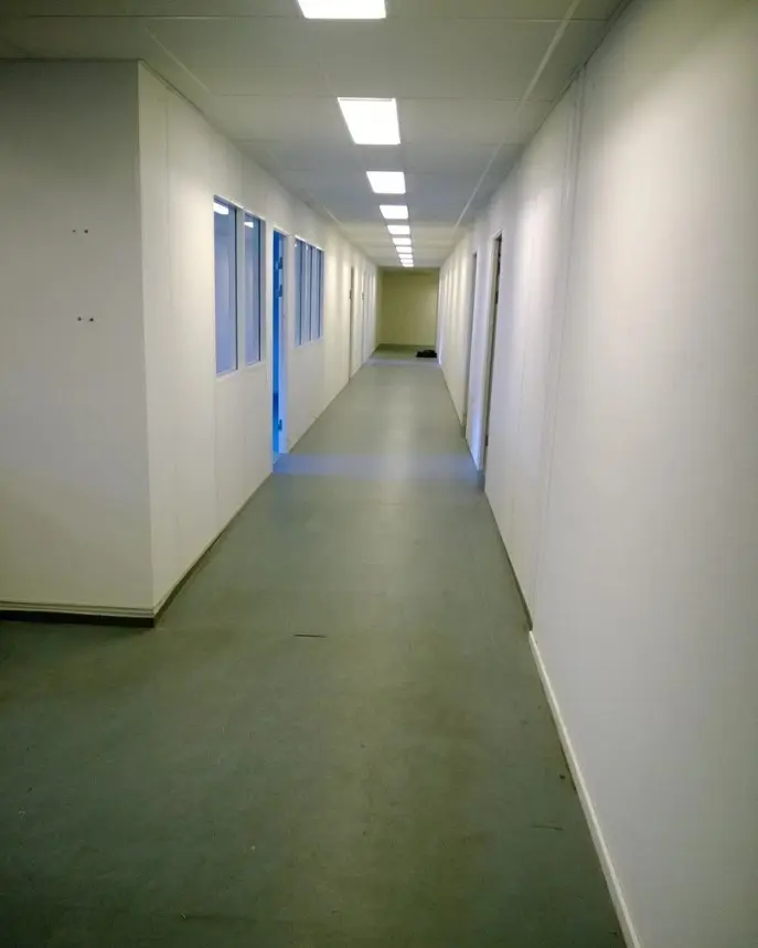 En lang, lys og tom gang i et kontor med hvide vægge og gule loftslamper. Gulvet er lavet af cement og er lysegråt