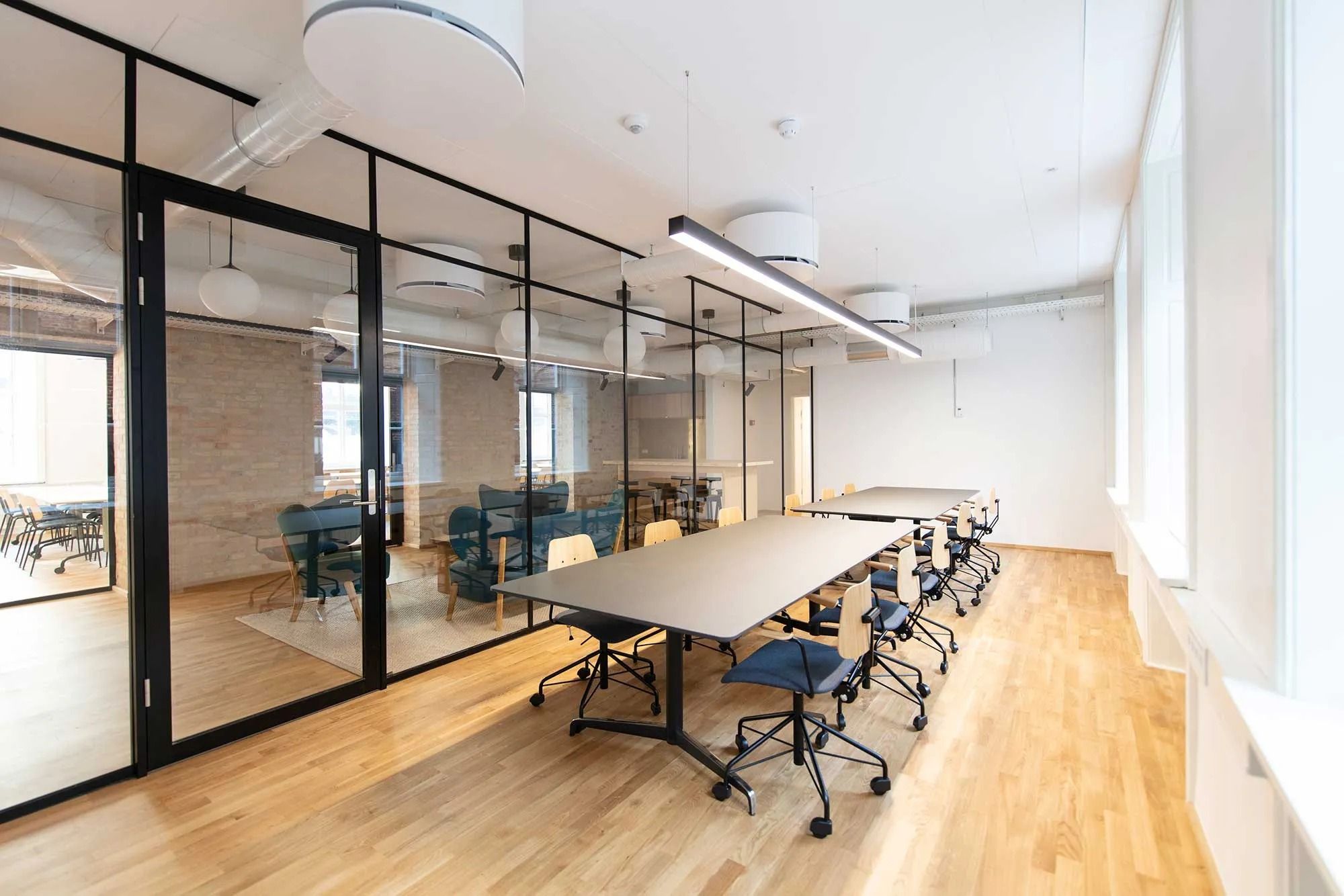 Minimalistisk mødelokale med trægulve og glasvægge, lysekroner hængende fra loftet, stolpeborde og stolpe, samt vinduer med