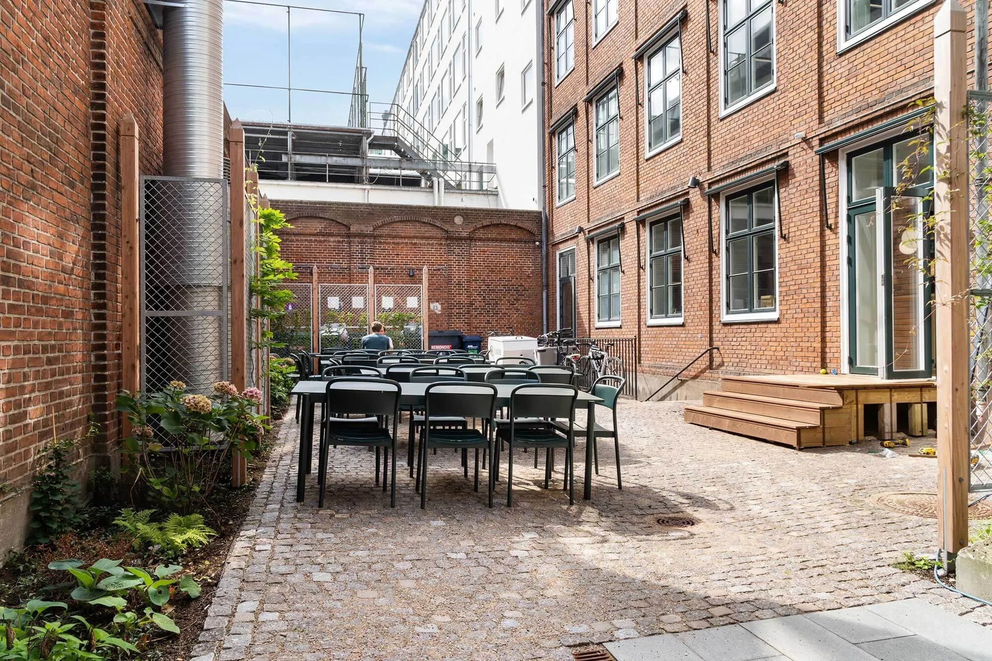 En gård med lyserøde blomster og grønne planter, flere stole og borde og en person i baggrunden, der står og ser mod venstre