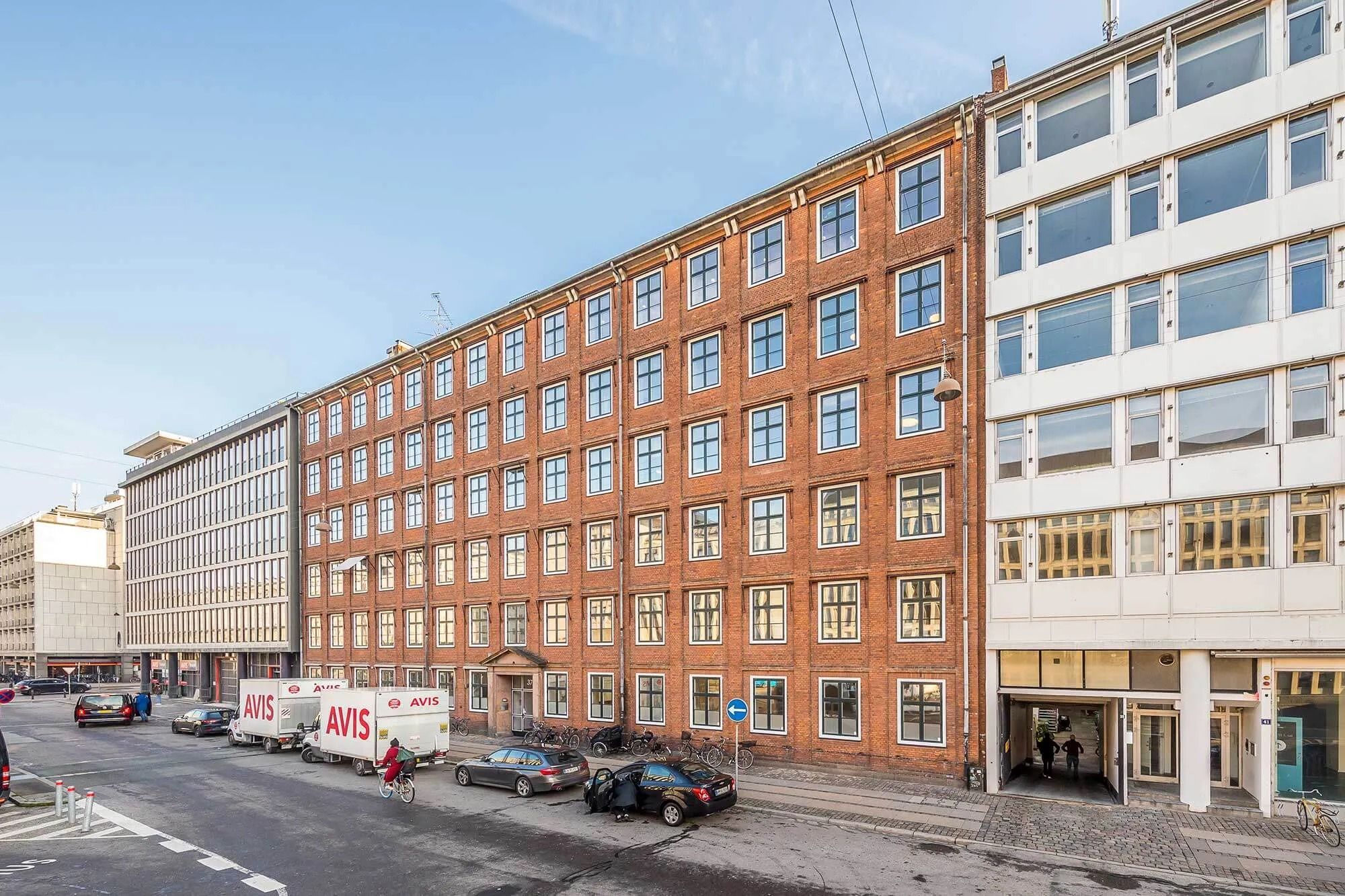 Avis-leverancebiler parkeret langs en gade med biler og cykelryttere, et stort kontorhus i baggrunden og en bygning med