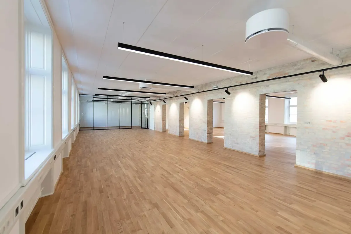 En stor, tom og lys kontorhall med højt til loftet, hvide vægge og trægulve, samt lysekroner og vinduer