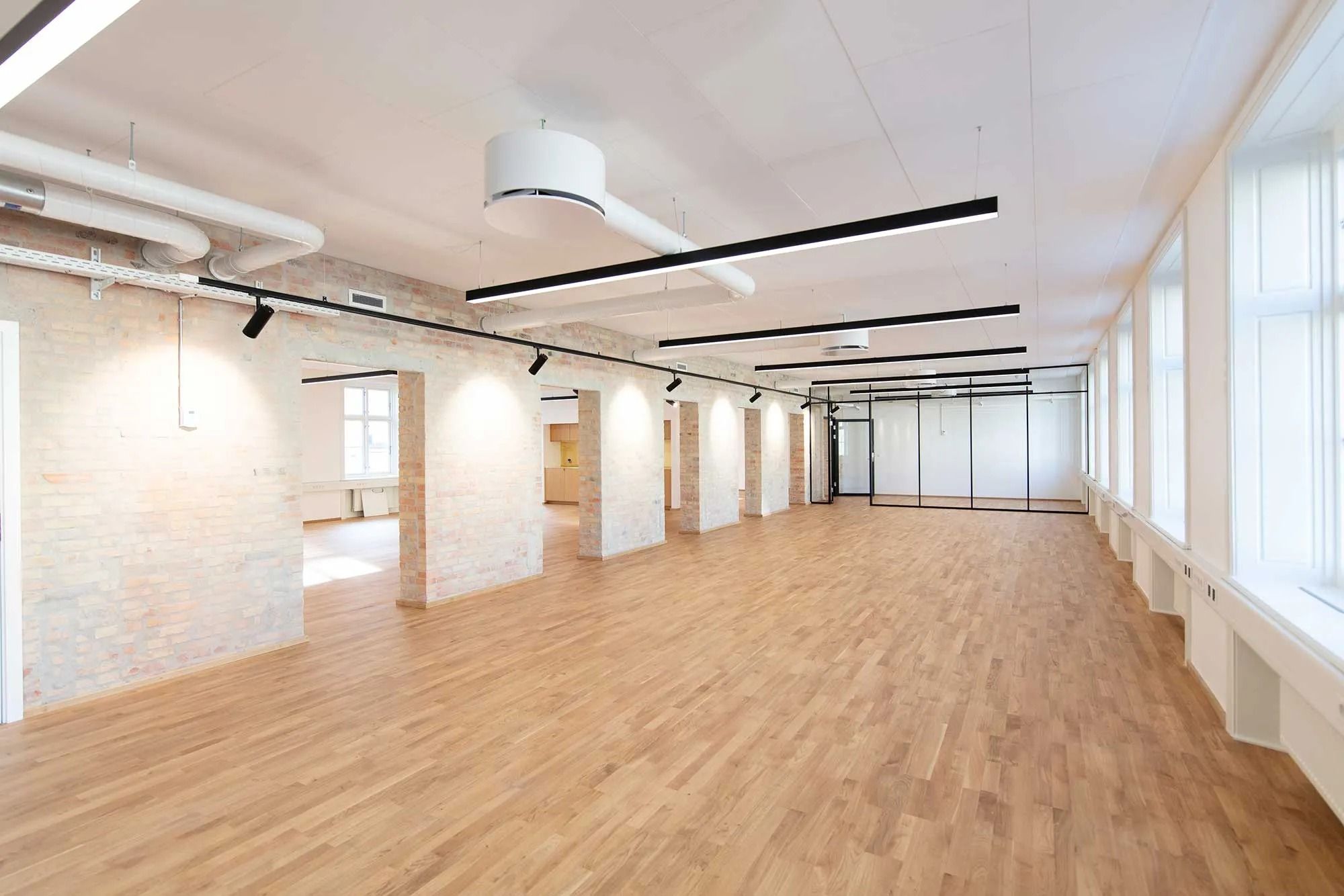 Tomt kontor med højt til loftet og store vinduer. Der er lyse trægulve og hvide vægge. To lysekroner hænger fra loftet og et