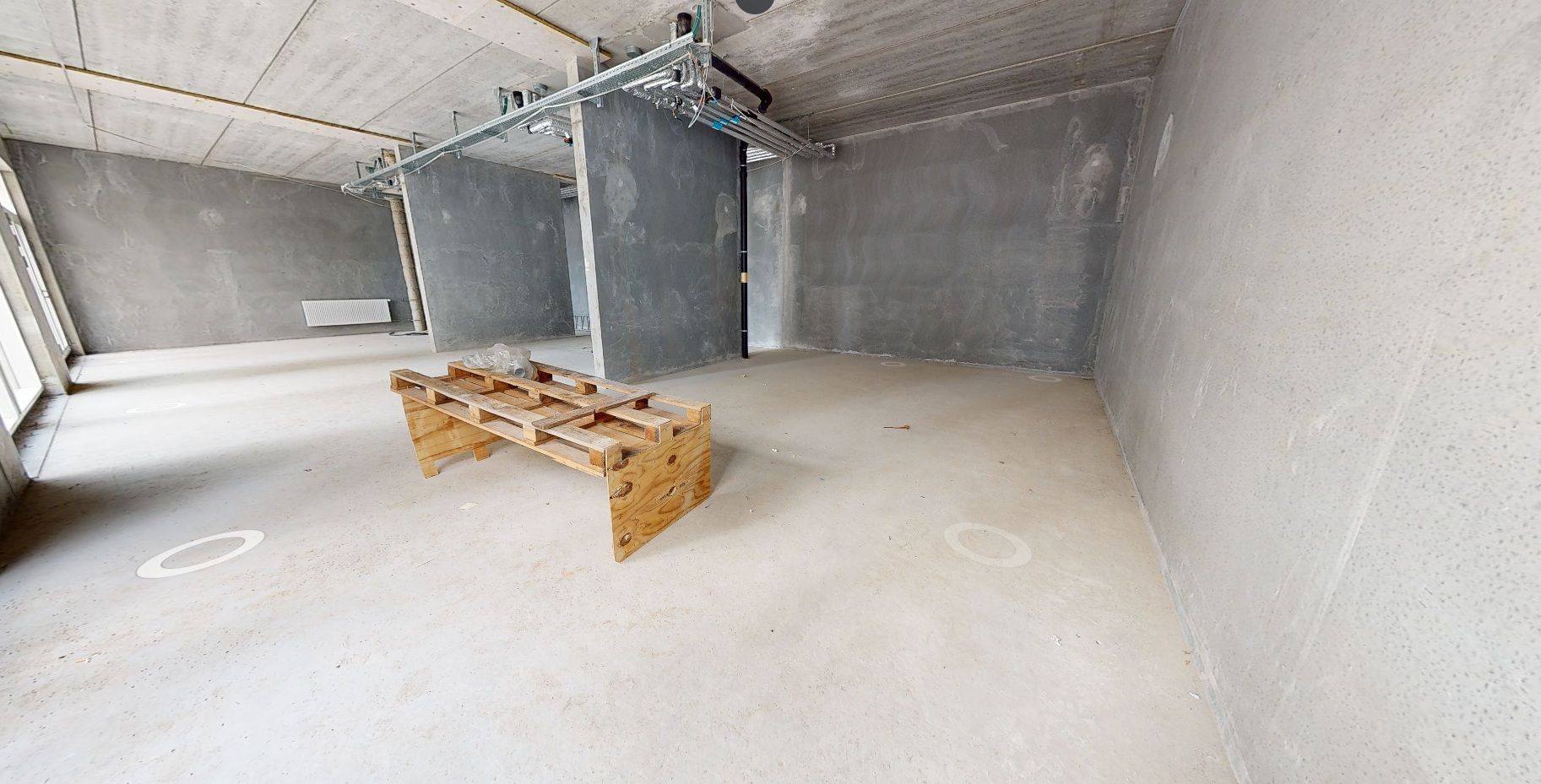 Et stort rum med hvide gulve og grå vægge med skabe og en træpalle. Vinduerne er åbne og der er et hvidt apparat på væggen.