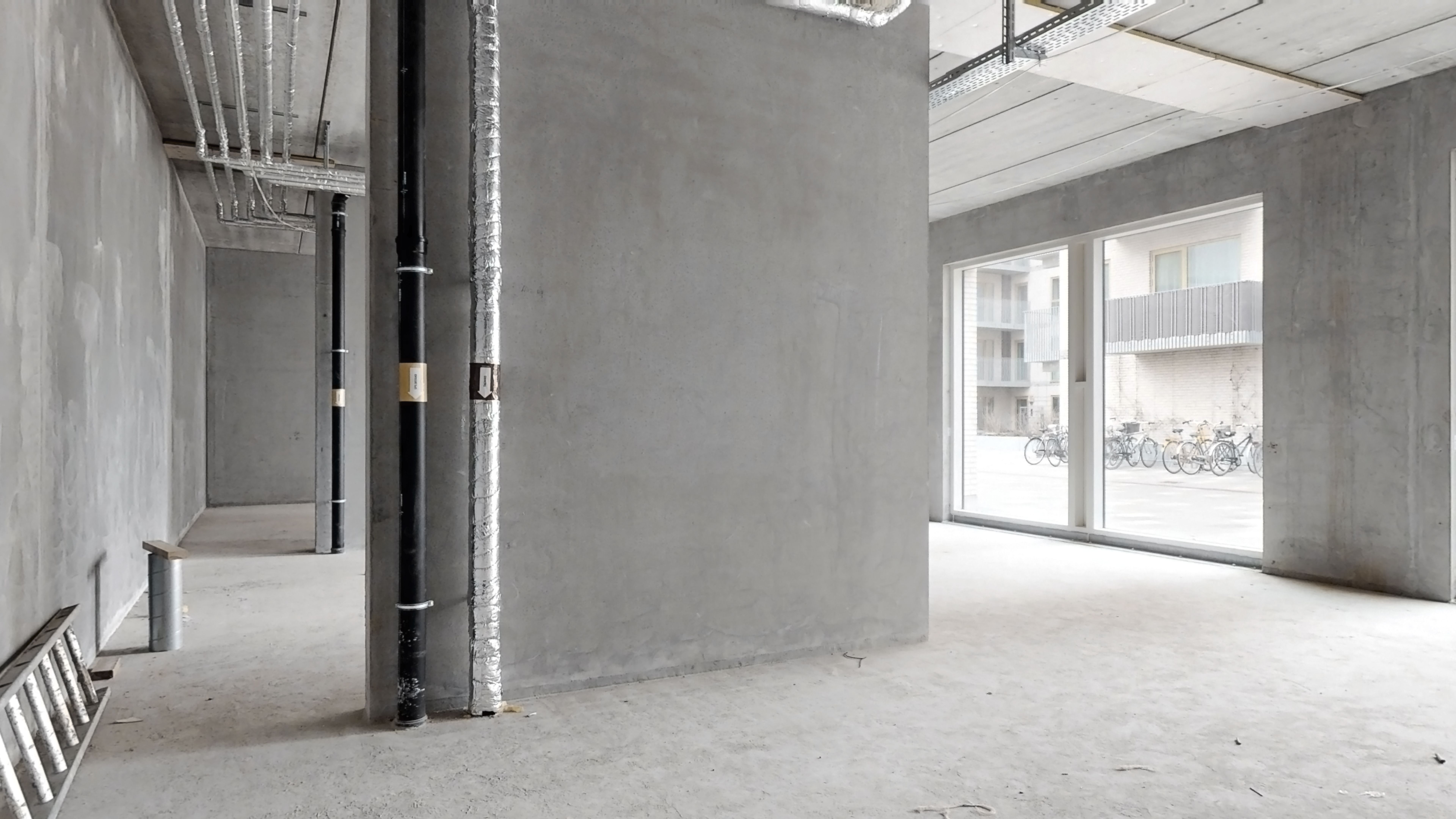 En stor bygning med højt loft og store ruder er i opførelse. Der er to bjælker og tre rør på væggen