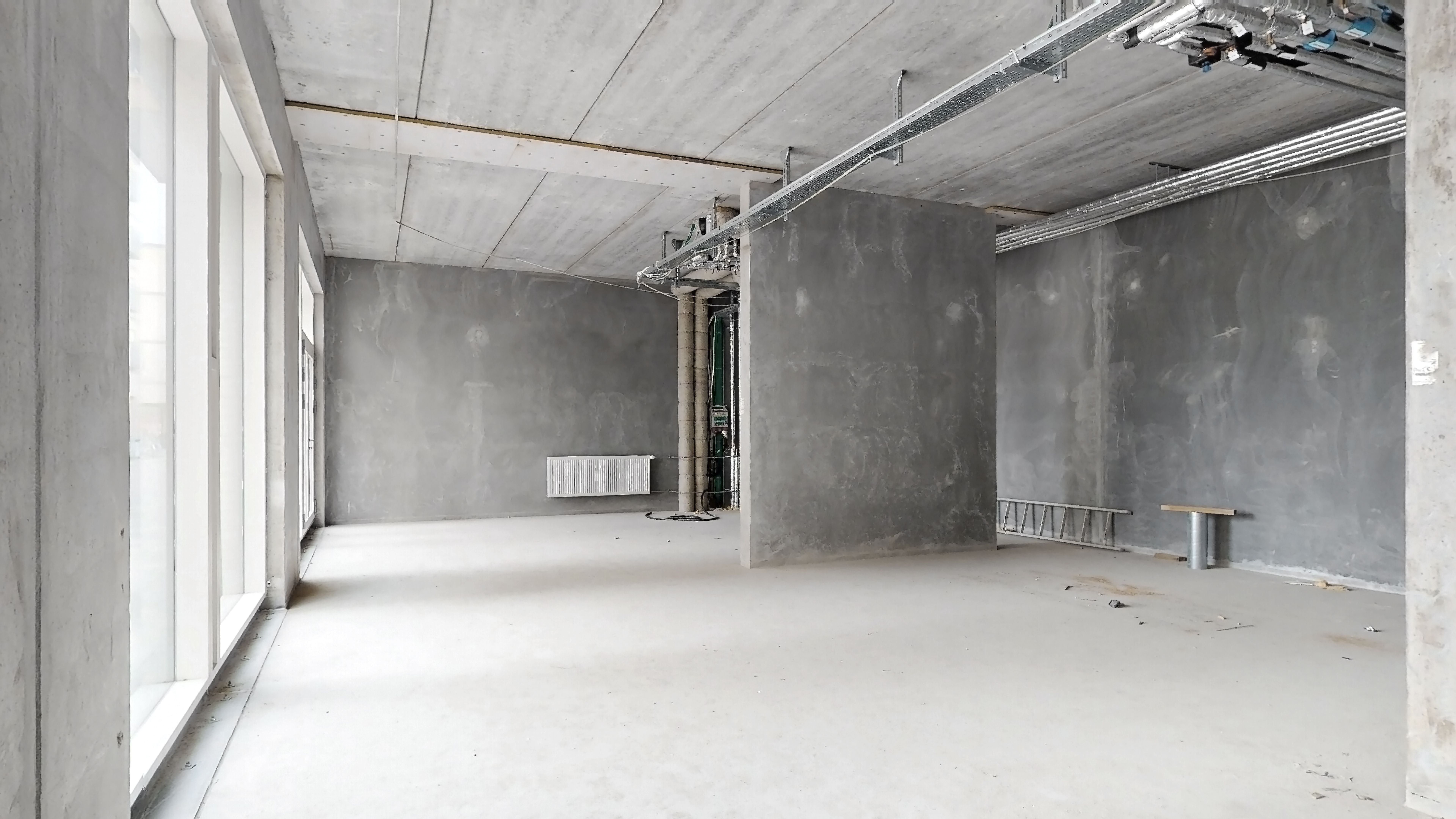 Det er en tom bygning med lysegrå vægge og et hvidt gulv. Der er et rødt gulv, et hvidt radiator, og et metalrør og en stige