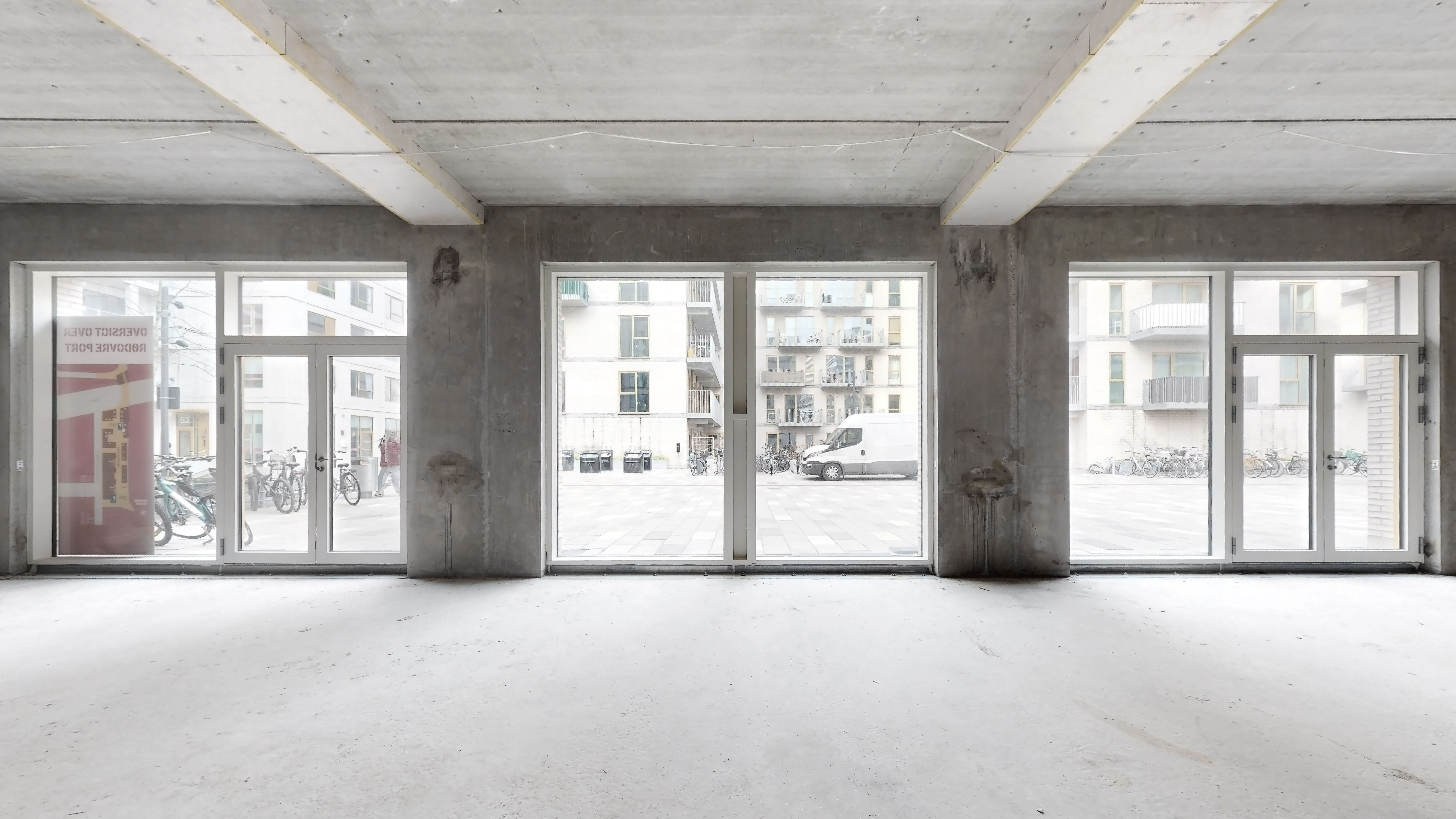 Rum med høje glasvinduer og hvidgulv og søjler med højtloft. Udsigt til en parkeringsplads med cykler og en hvid lastbil.