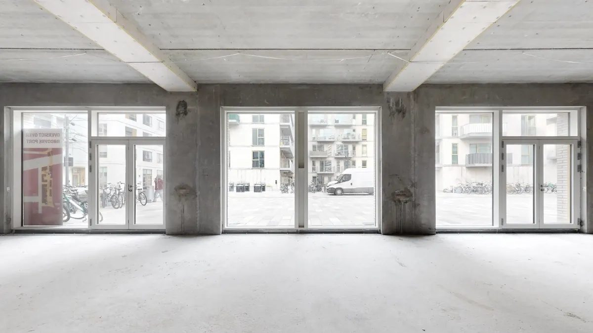 Rum med høje glasvinduer og hvidgulv og søjler med højtloft. Udsigt til en parkeringsplads med cykler og en hvid lastbil.