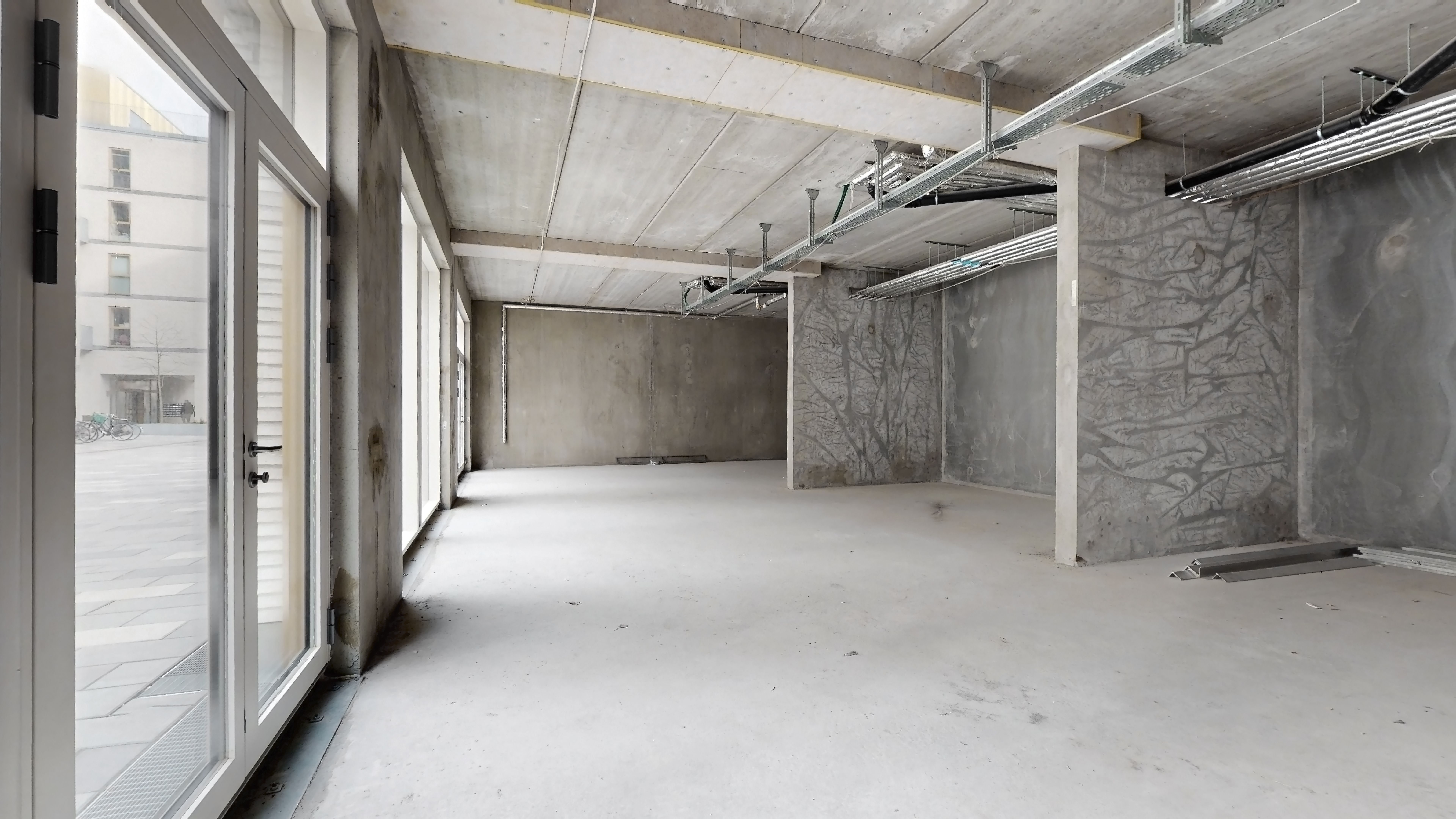 En ny, tom lejlighed med cementgulv og vinduer mod en gågade med cykler og bygninger i baggrunden