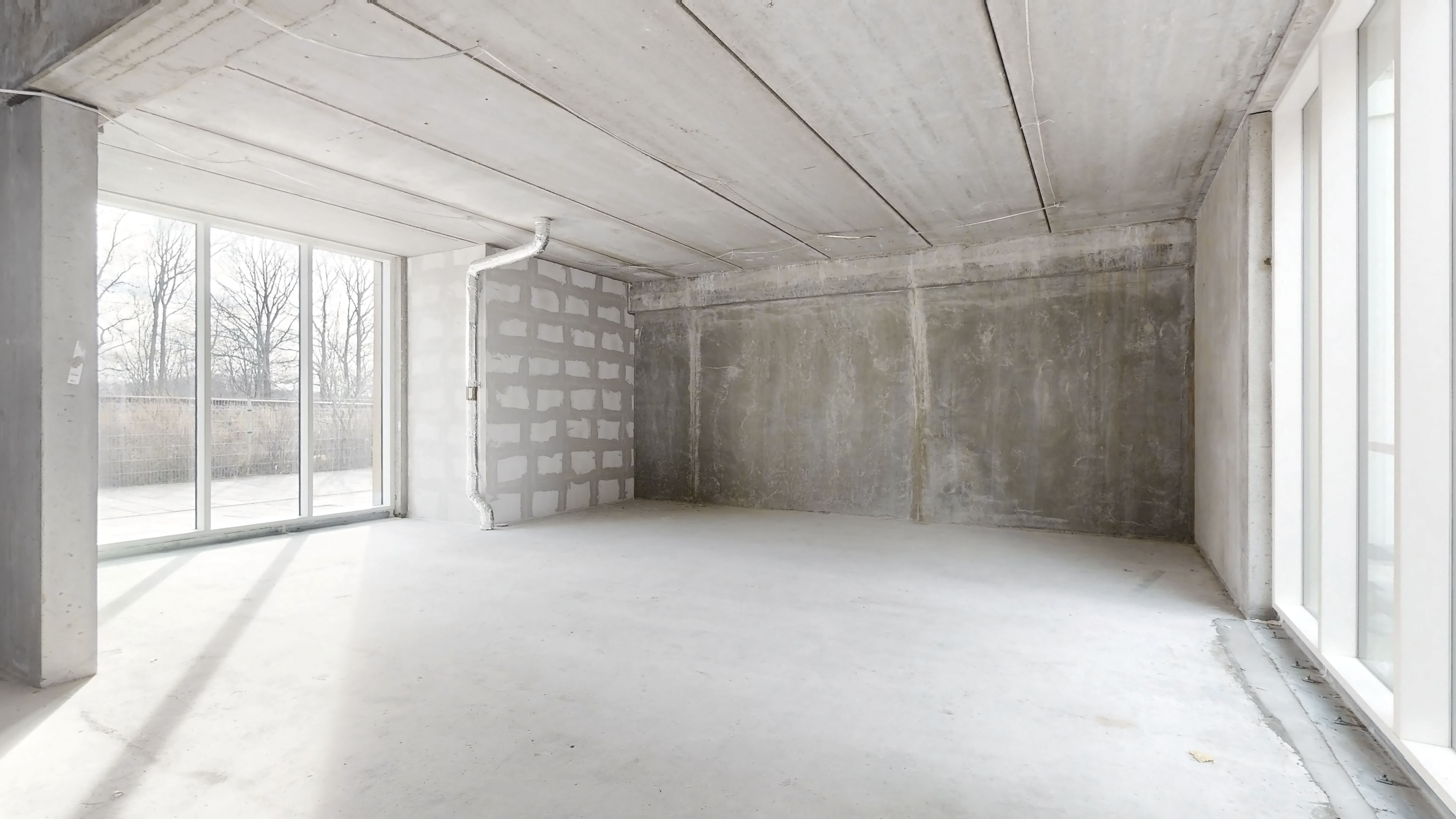 Et rum med hvide vægge og cementgulv og en stor glasdør med udsigt til træer og vej.