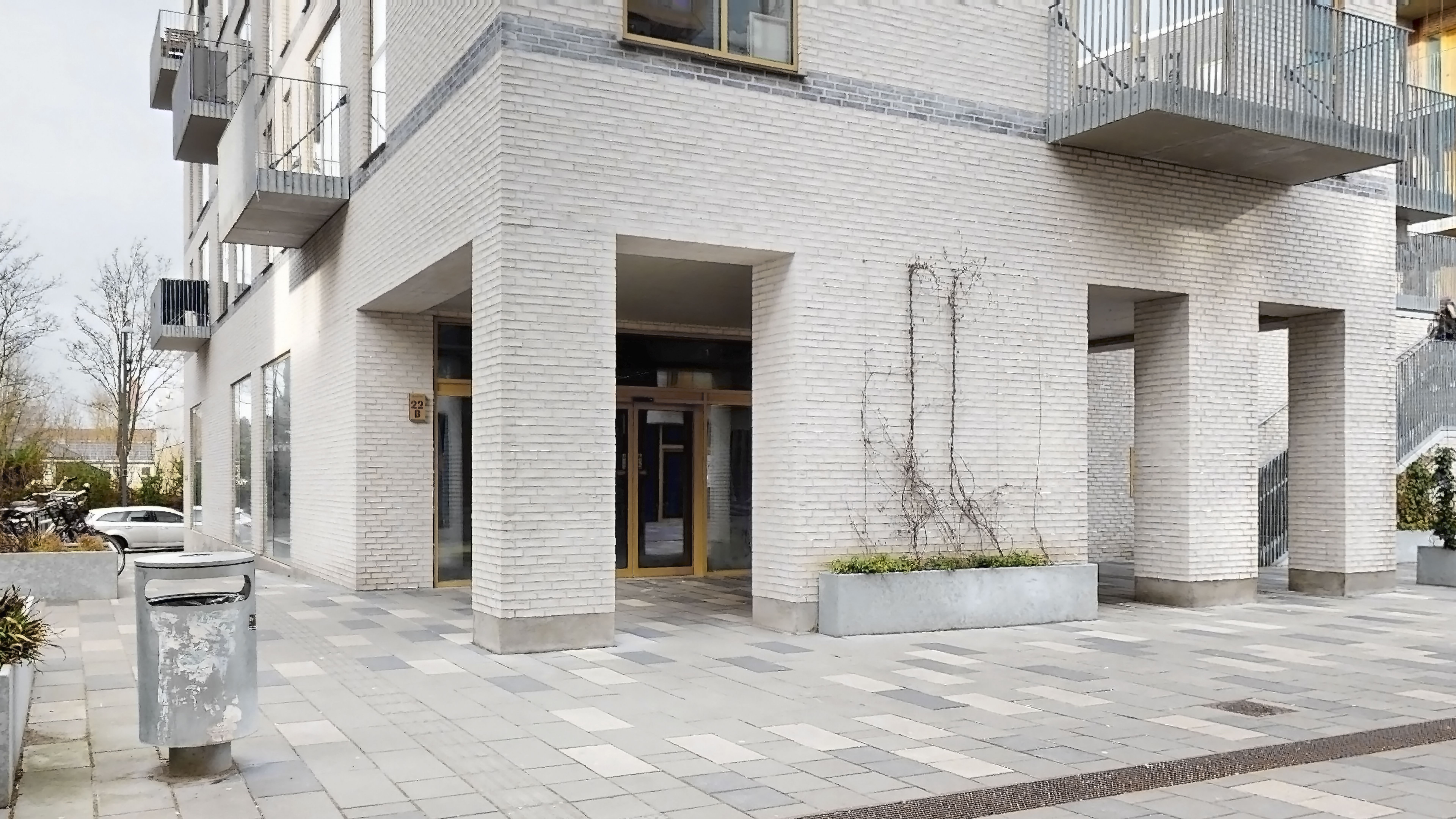 Hvid bygning med fire indgange til lejligheder. Træstammer og planter udenfor. Sted med gul tømmer og røde tage.