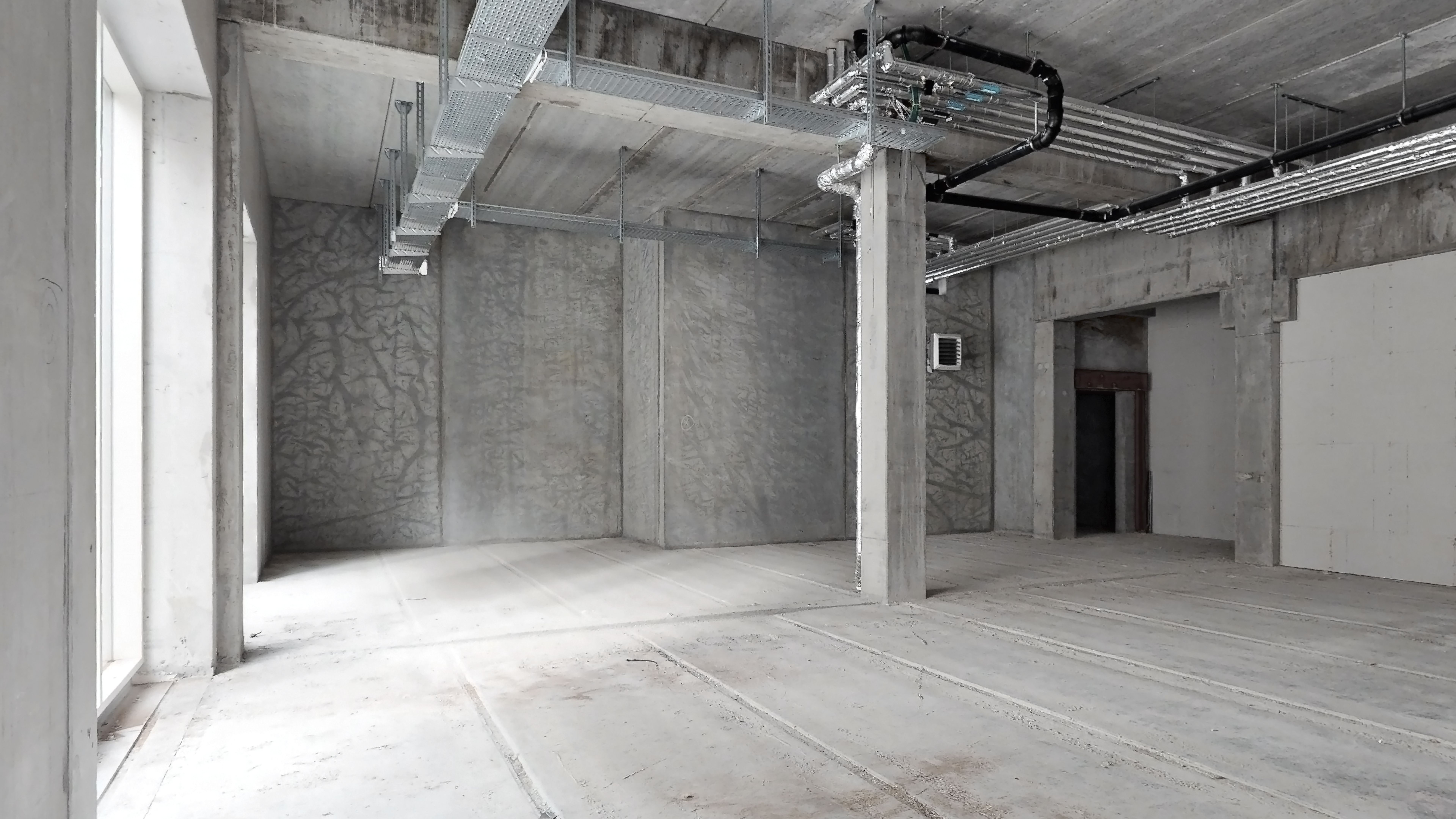 En stor, tom rum med lyse, teksturerede vægge og en høj, slank søjle samt flere luftkanaler og rør ophængt i loftet