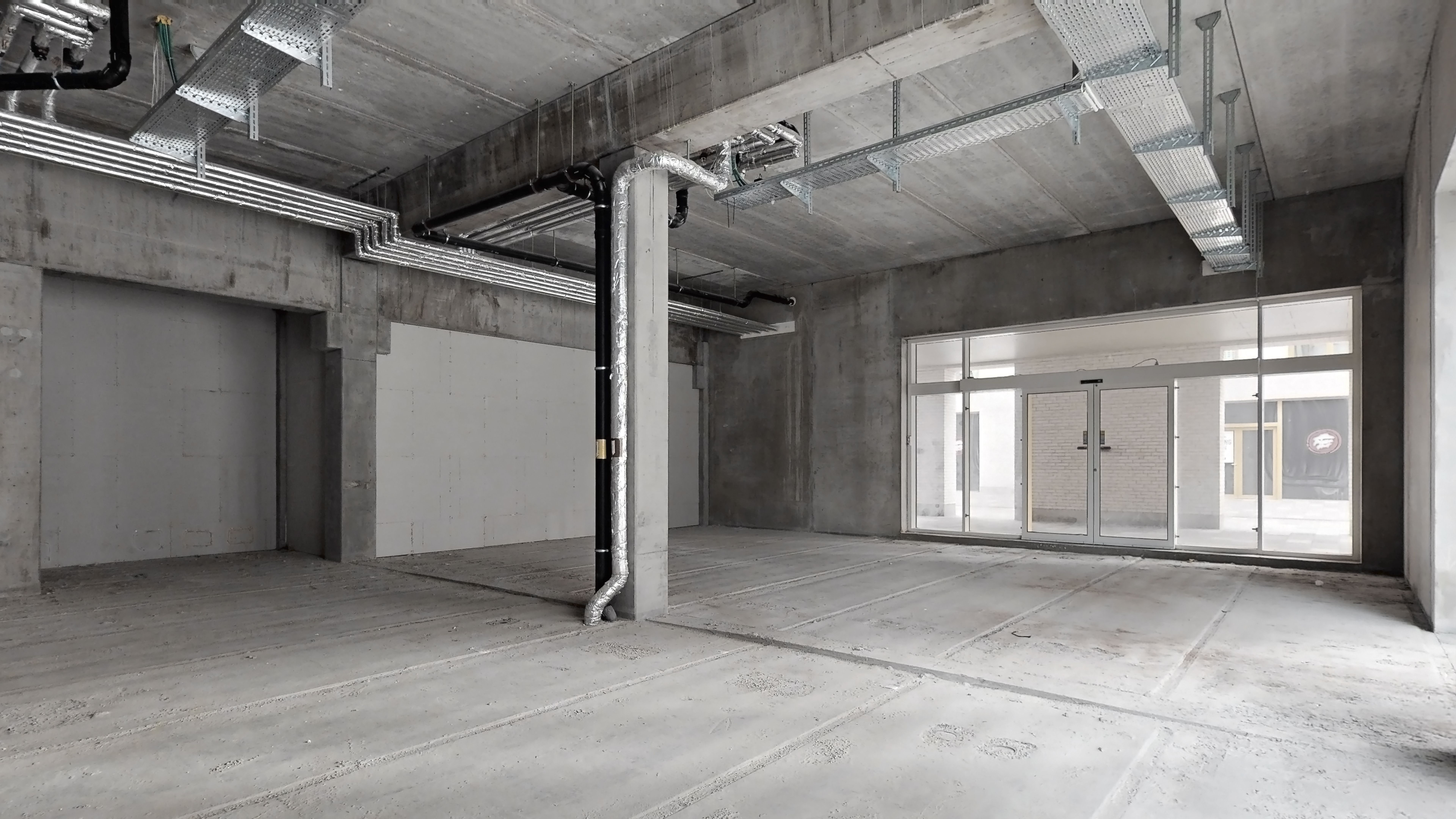 Et stort rum med høje lofter, en hvid væg og glasdøre. Der er en metalkonstruktion og en søjle med røgledning. Gulvet er