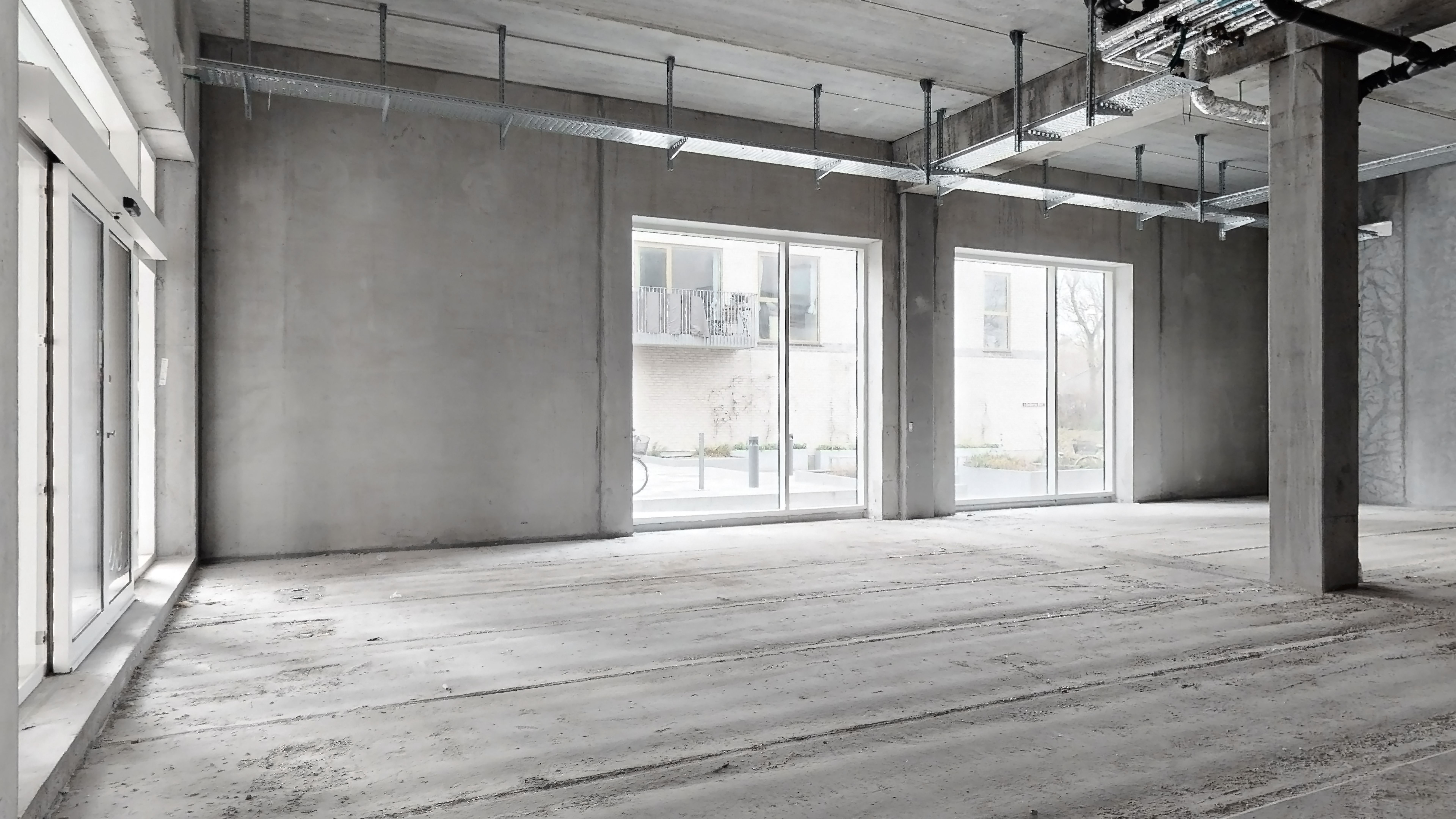 Betongulv og vægge i en stor, lys hall med glasdøre og vinduer med udsigt til grønne haver og en balkon