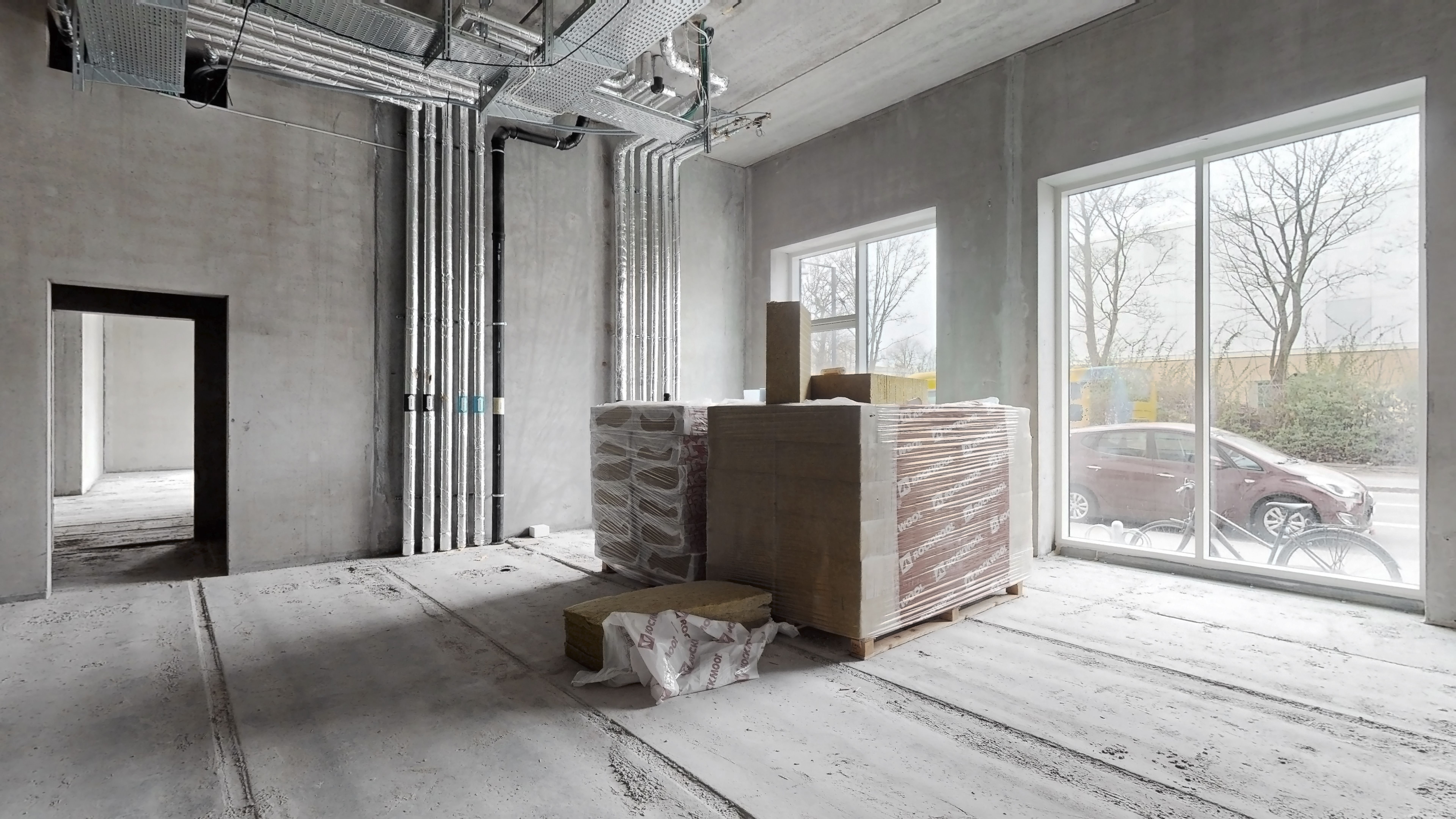 Indvendigt i en nyopført bygning med vinduer og åbne døre. Byggematerialer og emballage er placeret på gulvet. Udvendigt er
