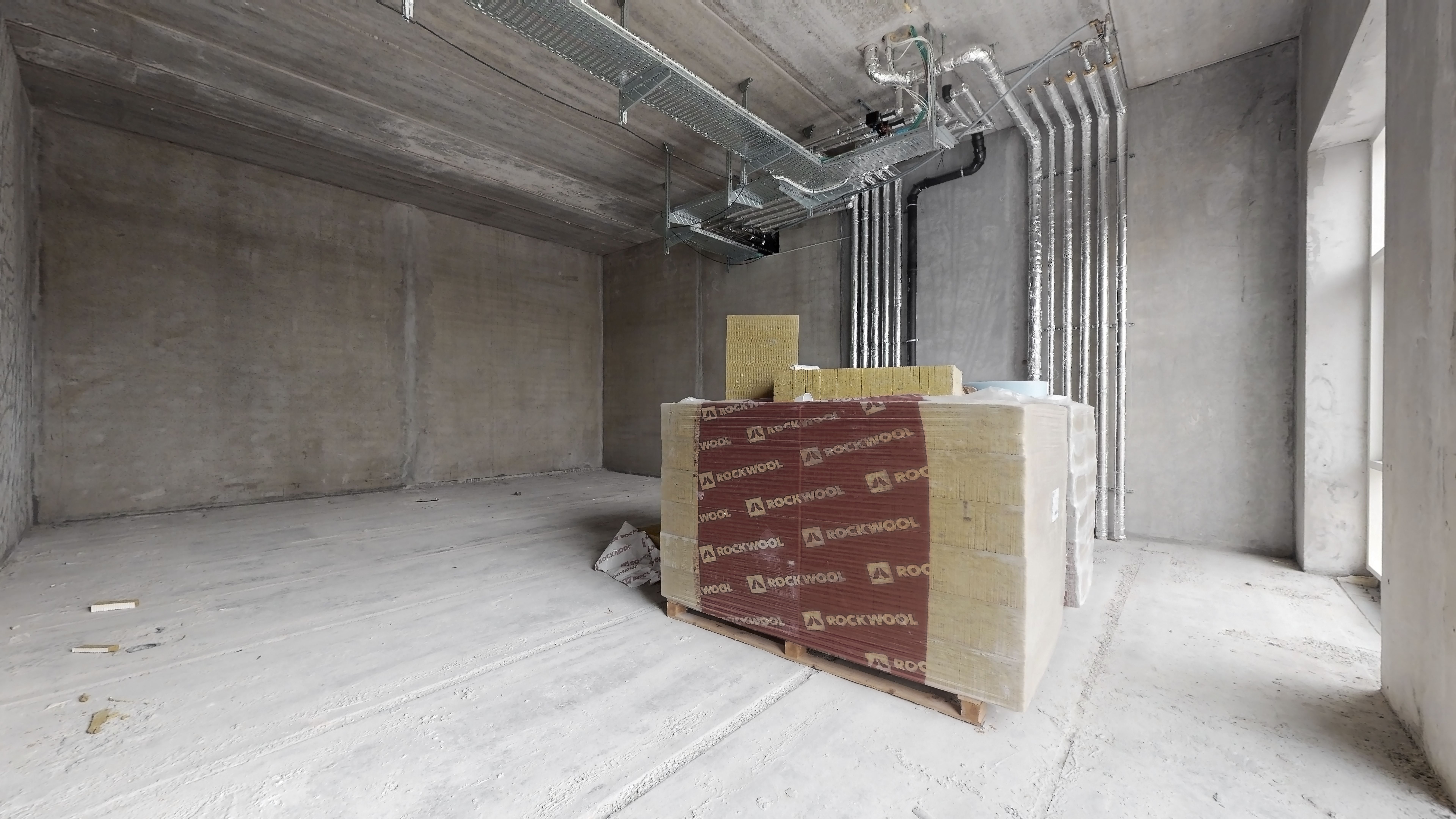 Beton vægge og loft samt en række varmelegemer og rør. En stor kasse med Rockwool og andet materiale står på en palle