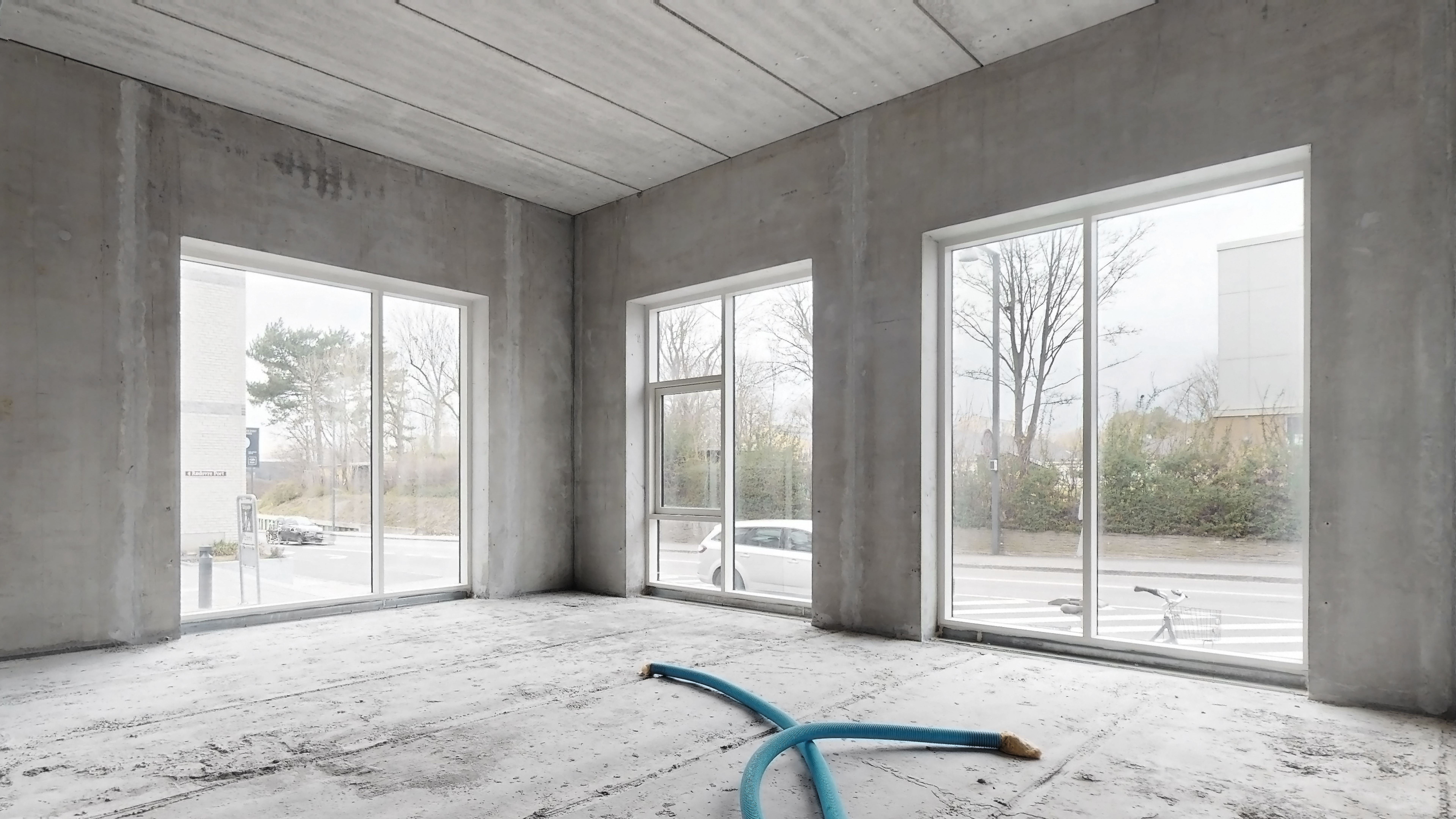 Bygget rum med trægulv og store vinduer mod vejen udenfor med biler og træer og uden for vinduet er der en cykel