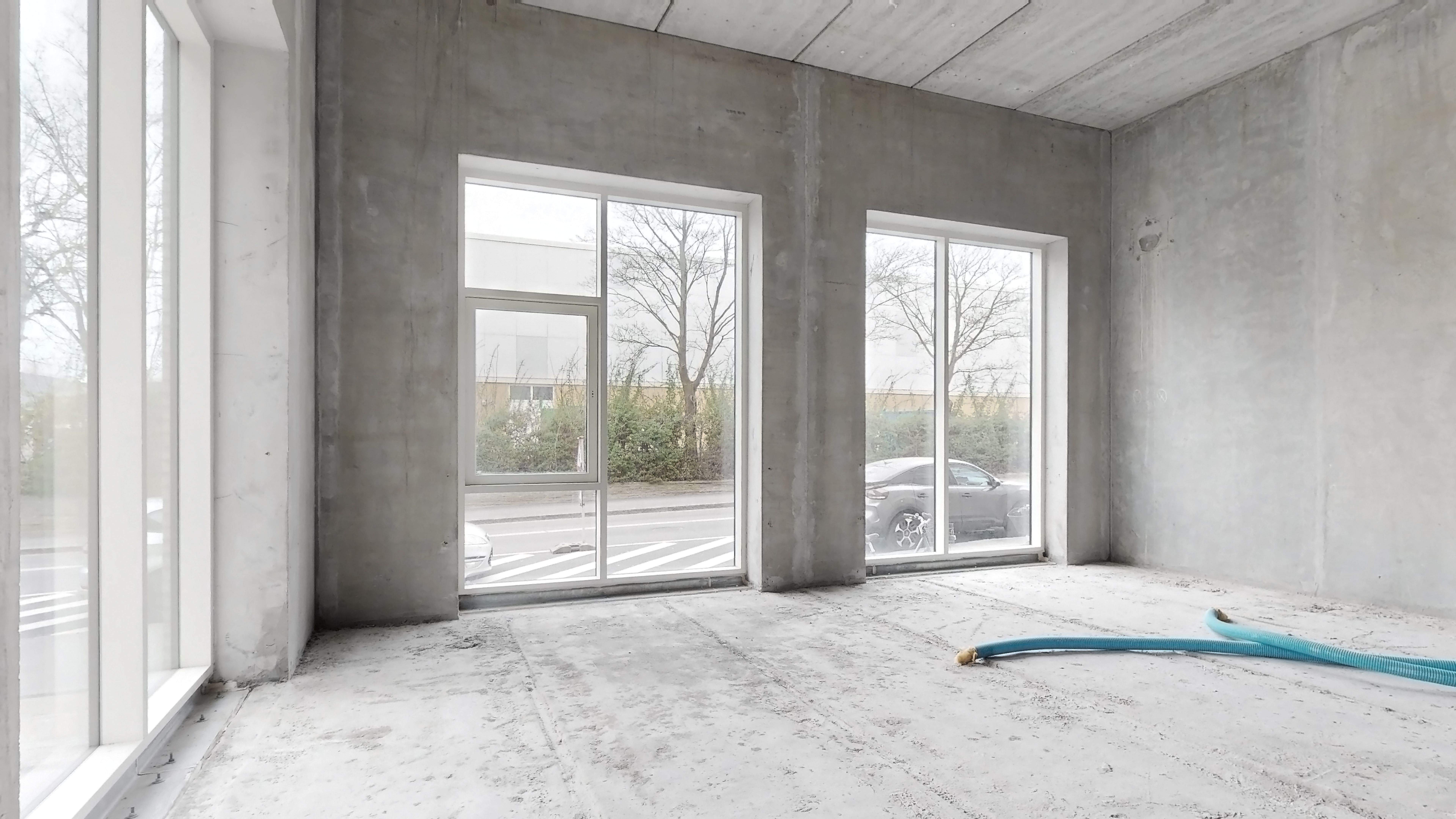Et tomt værelse med glasvinduer og døre, cementgulv, og en slange på gulvet. En bil parkeret på gaden udenfor, og træer og