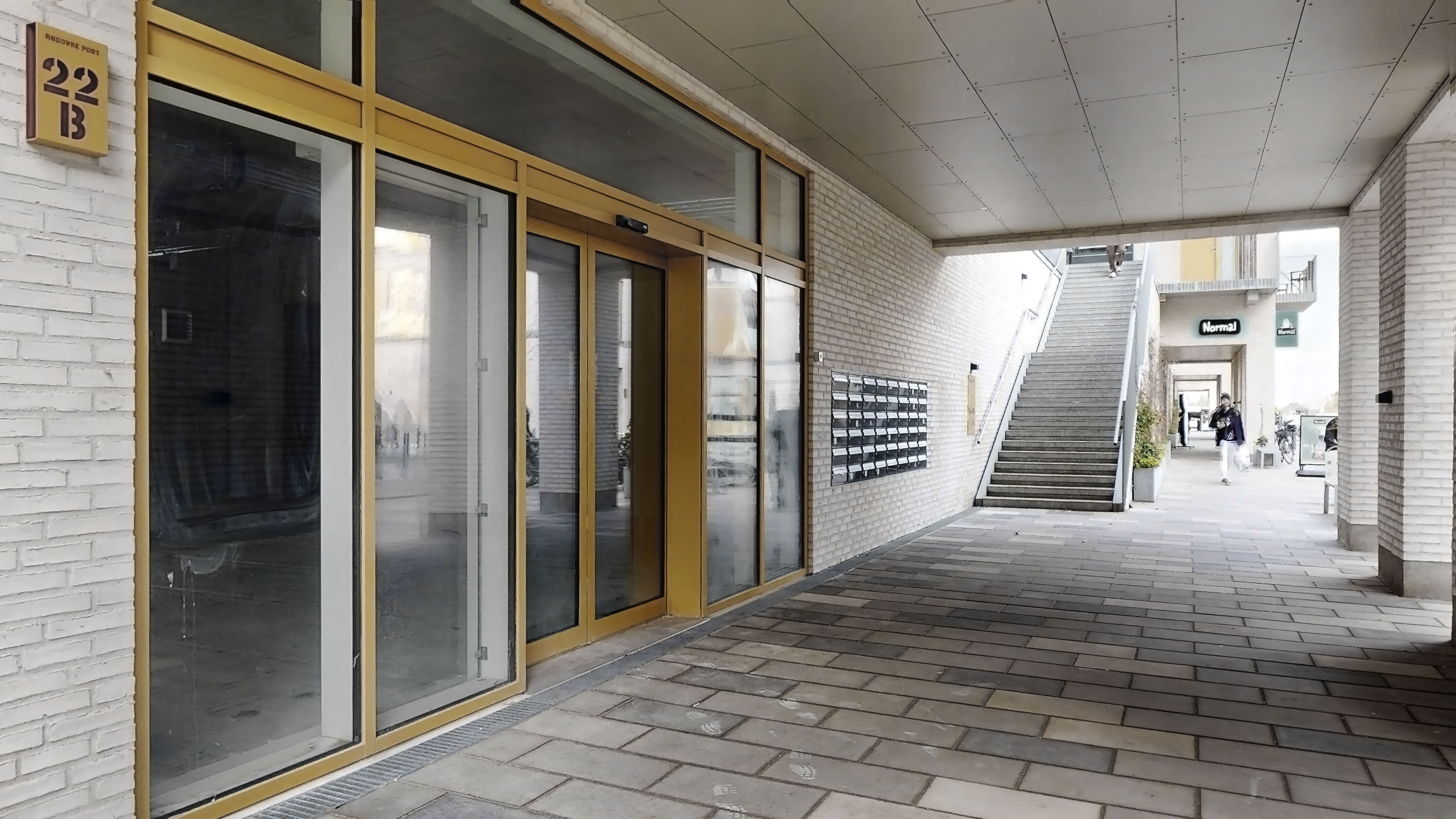 Et glasdækket indgangsparti med nummer 22B, udstyret med moderne gule døre og gulv af mørke mursten, der fører til et