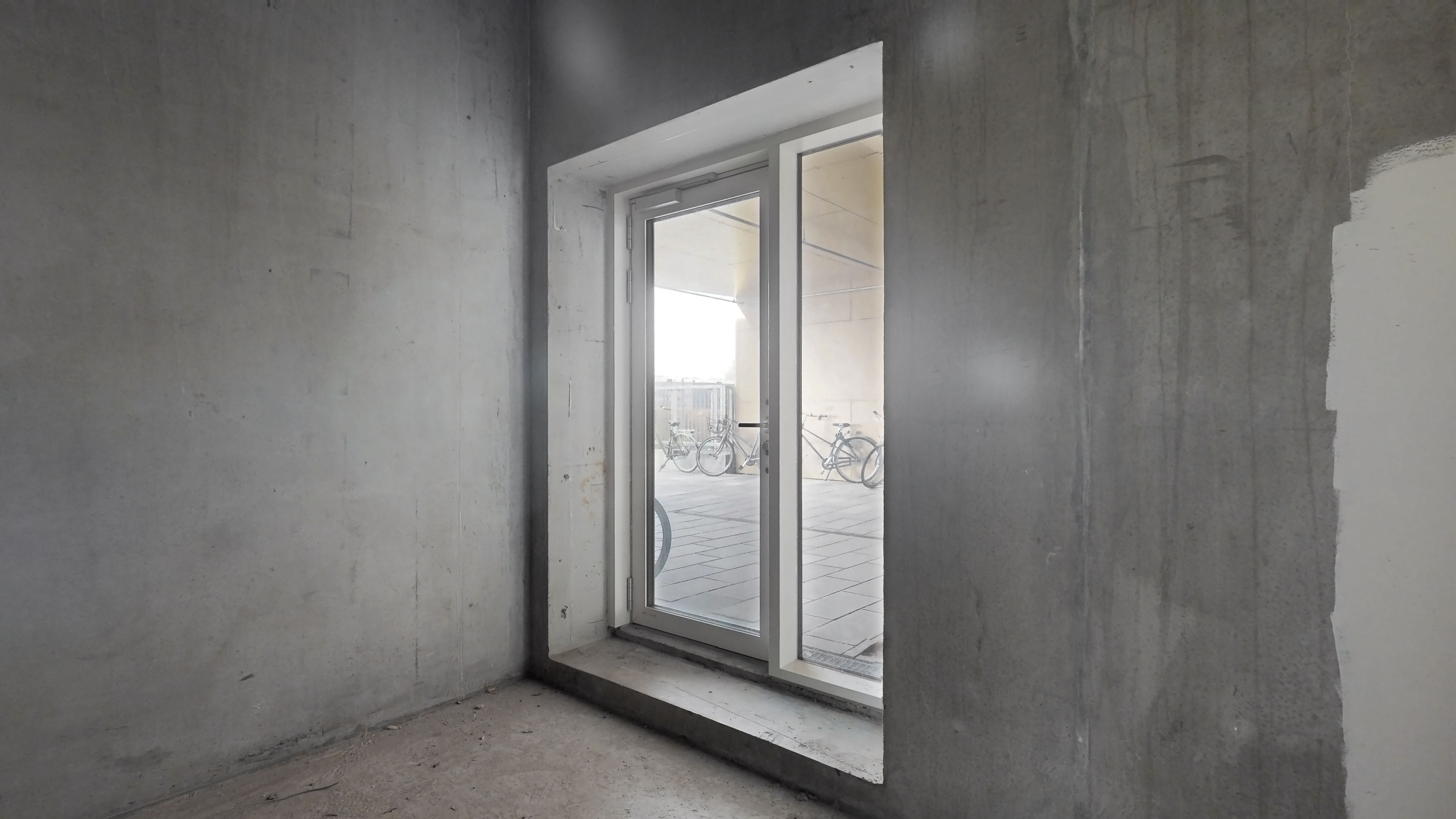 Et skarpt billede af et tomt værelse med cementvægge og et stort glasvindue, der vender ud mod en terrasse med cykler