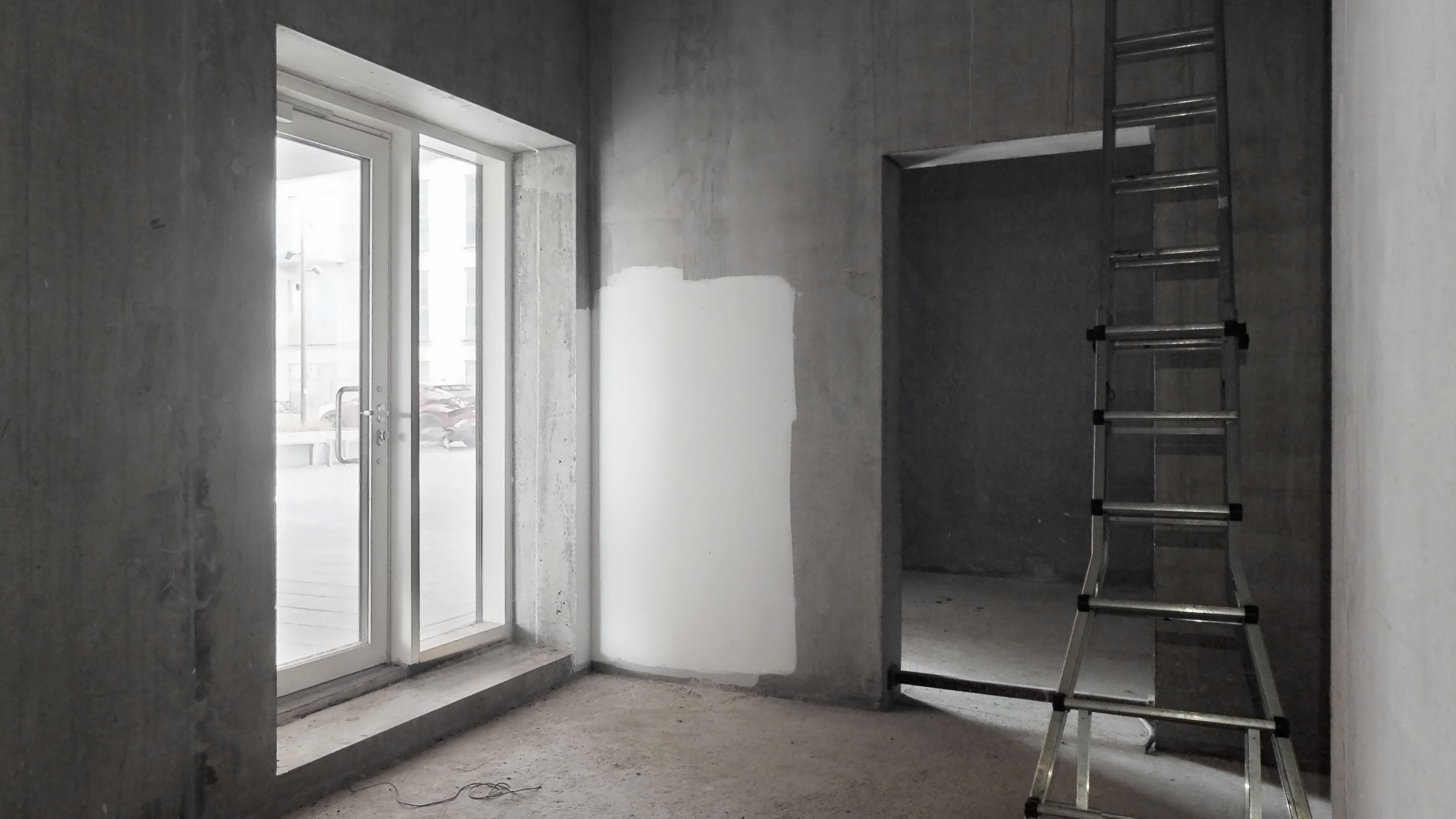 Byggepladsen med høje, grå vægge og vinduer med glasdøre, hvor der er en stige og en væg med ny gulvflise.
