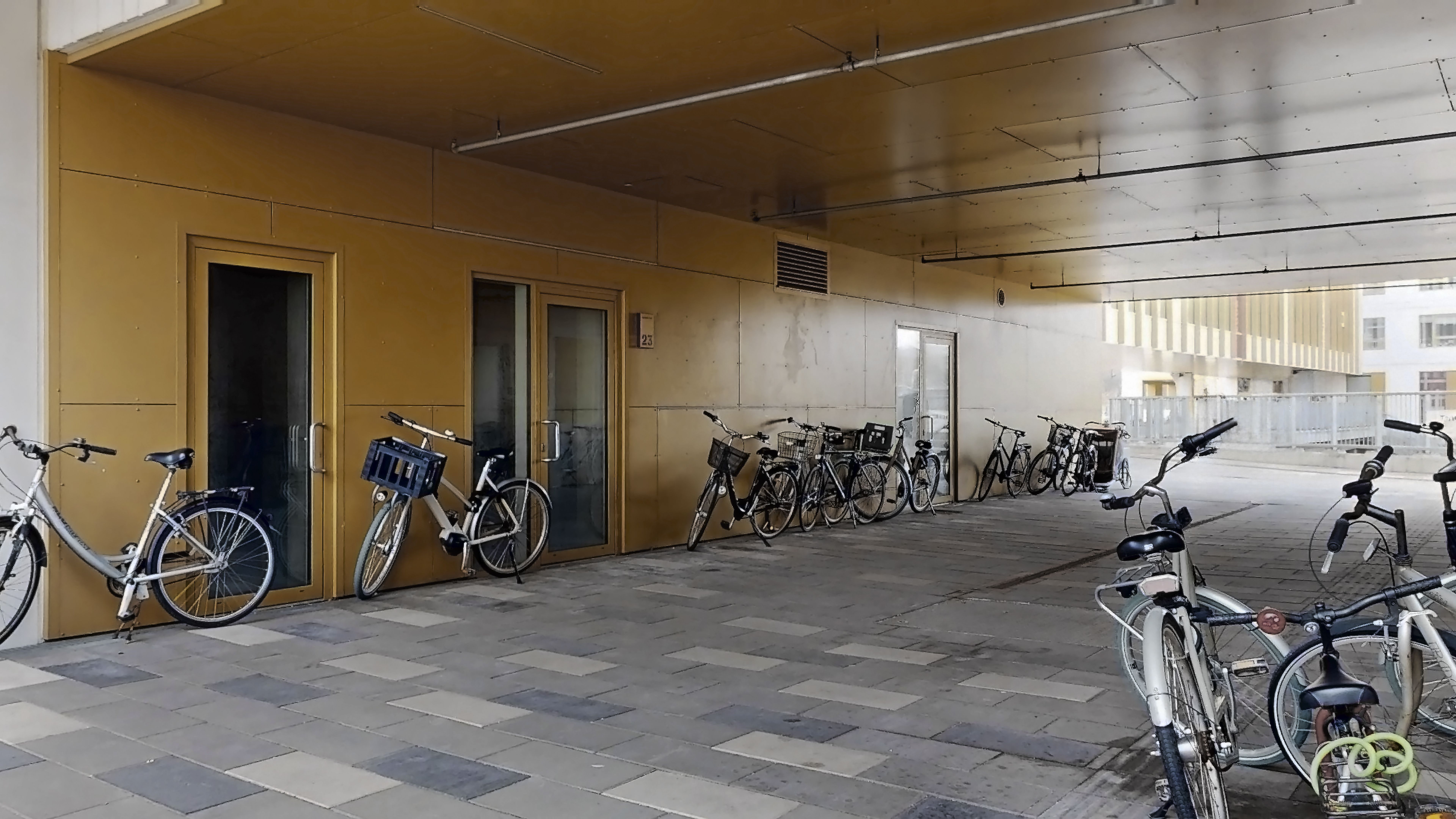 Parkerede cykler med kurve i en underjordisk cykelparkering, gulvbelægning i kryds og to gule døre med nummerplade 23
