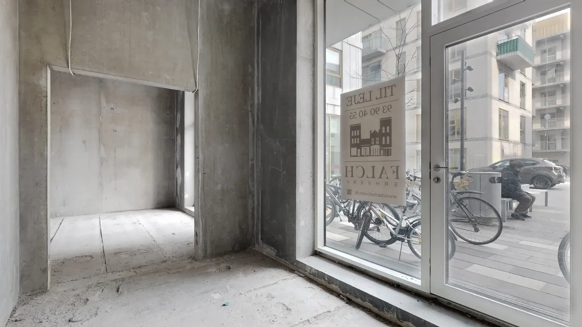 Udstødet lokale i en ny bydel i Rødovre med et skilt, der siger "Flat for rent". På gaden udenfor parkerer biler og cykler,