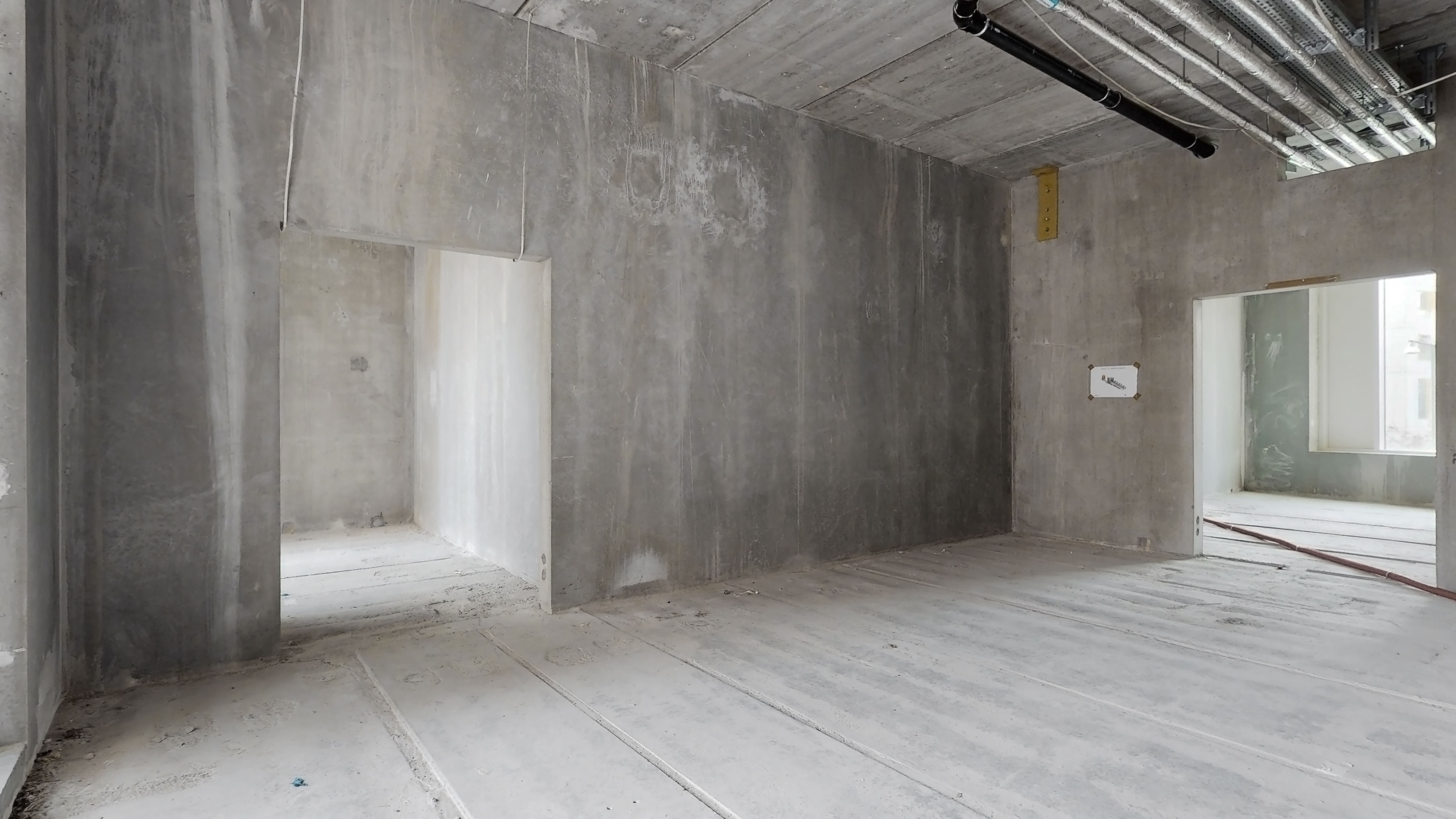 En stor, tom rum med højt loft og betonvægge, to døre og en vinduesåbning samt rør og ledninger i loftet