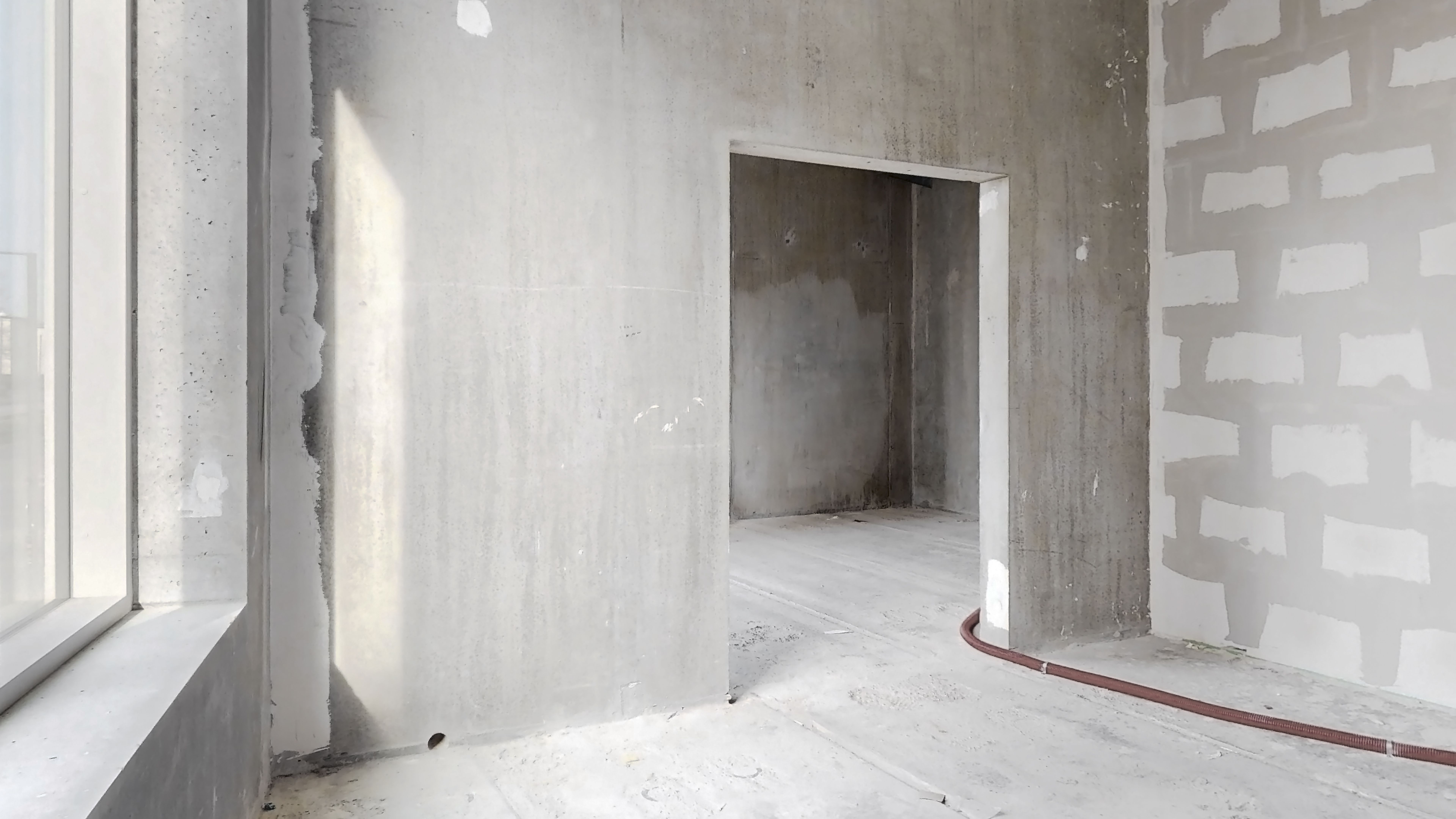 En ubeboet rum med hvide vægge og en åbning til en anden rum. En rør er liggende på gulvet og der er et lyskast på den ene