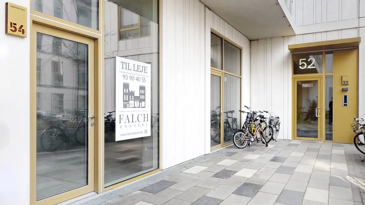 En lille gade med nummererede døre og parkerede cykler, hvor dør 54 har et skilt med teksten "TIL LEJE" og dør 52 har et