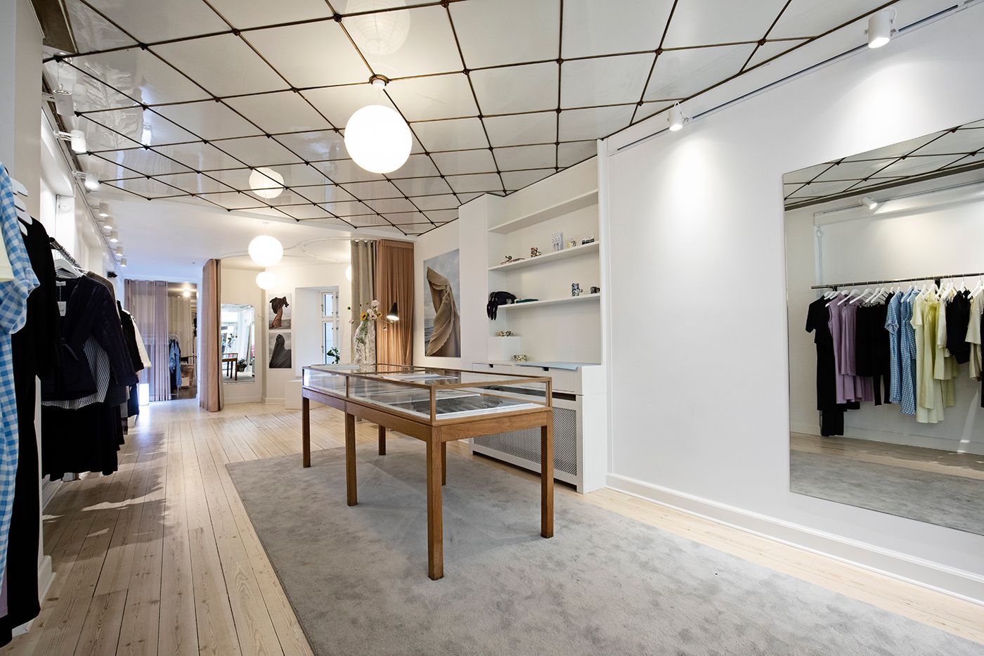 Luksuriøs tøjbutik med lyse trægulve og moderne indretning, hvid væg og spejl, og tøj på klædhængere samt en stol og et bord